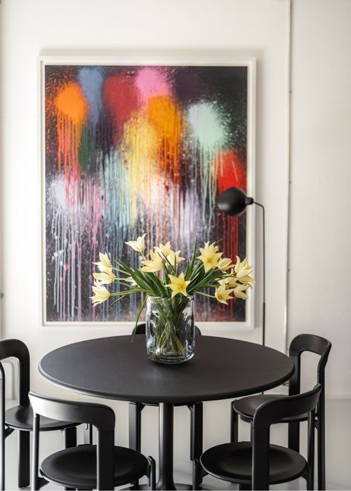Ein runder schwarzer Tisch mit passenden Stühlen. Auf dem Tisch befindet sich eine Vase mit frischen, gelben Blumen, die einen lebhaften Farbtupfer geben. Über dem Tisch hängt ein farbenfrohes Gemälde mit dynamischen, abstrakten Formen, das eine künstlerische Atmosphäre schafft.