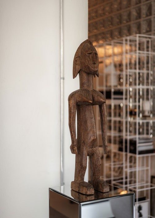 Eine rustikale Holzskulptur mit abstrakten Zügen steht auf einem modernen, spiegelnden Podest. Die Figur, anscheinend inspiriert von tribaler Kunst, verleiht dem modernen Raum eine Note von Tradition und Geschichte.