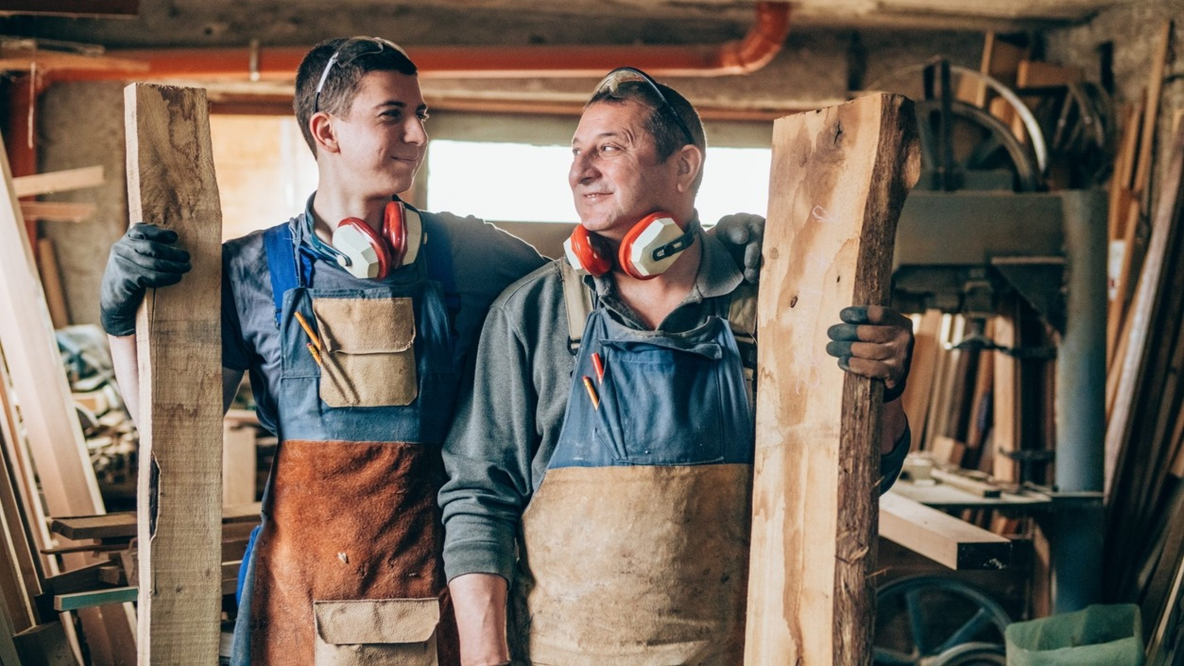 Deux collaborateurs de générations différentes sont dans un atelier et tiennent chacun une poutre en bois.