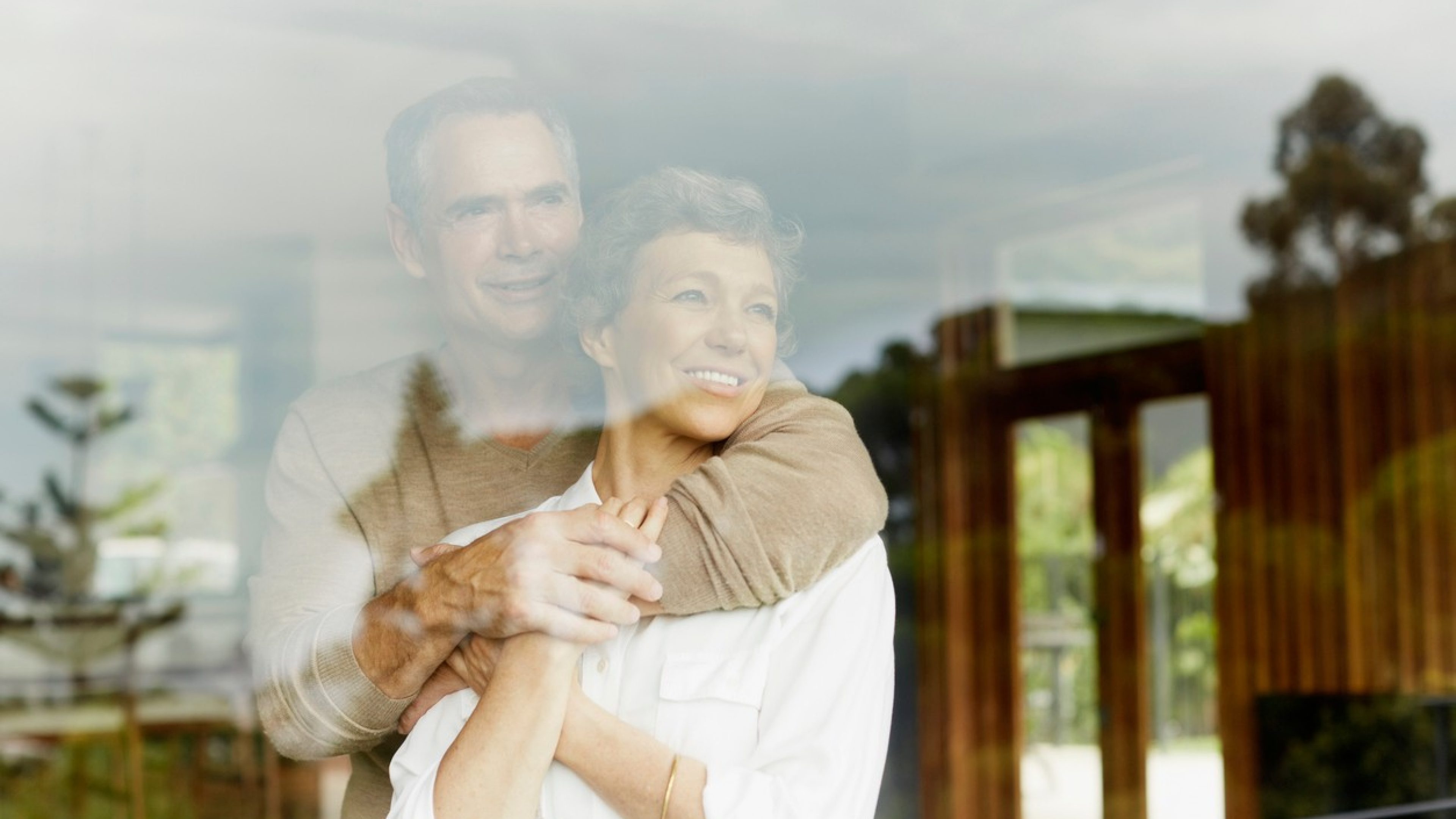 Una coppia over 50 si abbraccia e guarda fuori dalla finestra. I colori sono caldi. L’uomo indossa un maglione beige, la donna una camicetta bianca. Entrambi sorridono e guardano in lontananza dalla finestra.