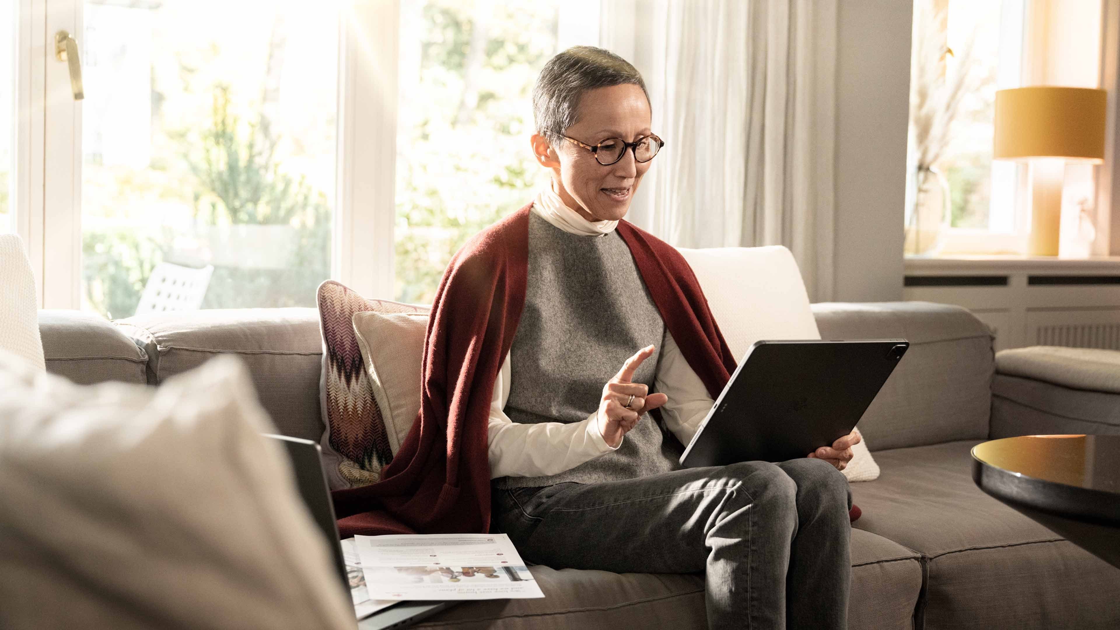 Eine ältere weibliche Person sitzt mit einem geöffneten Notebook auf einem Sofa. Neben ihr liegen diverse Dokumente auf einem Papierstapel. Sie studiert den Screen und hat ihre rechte Hand leicht erhoben.