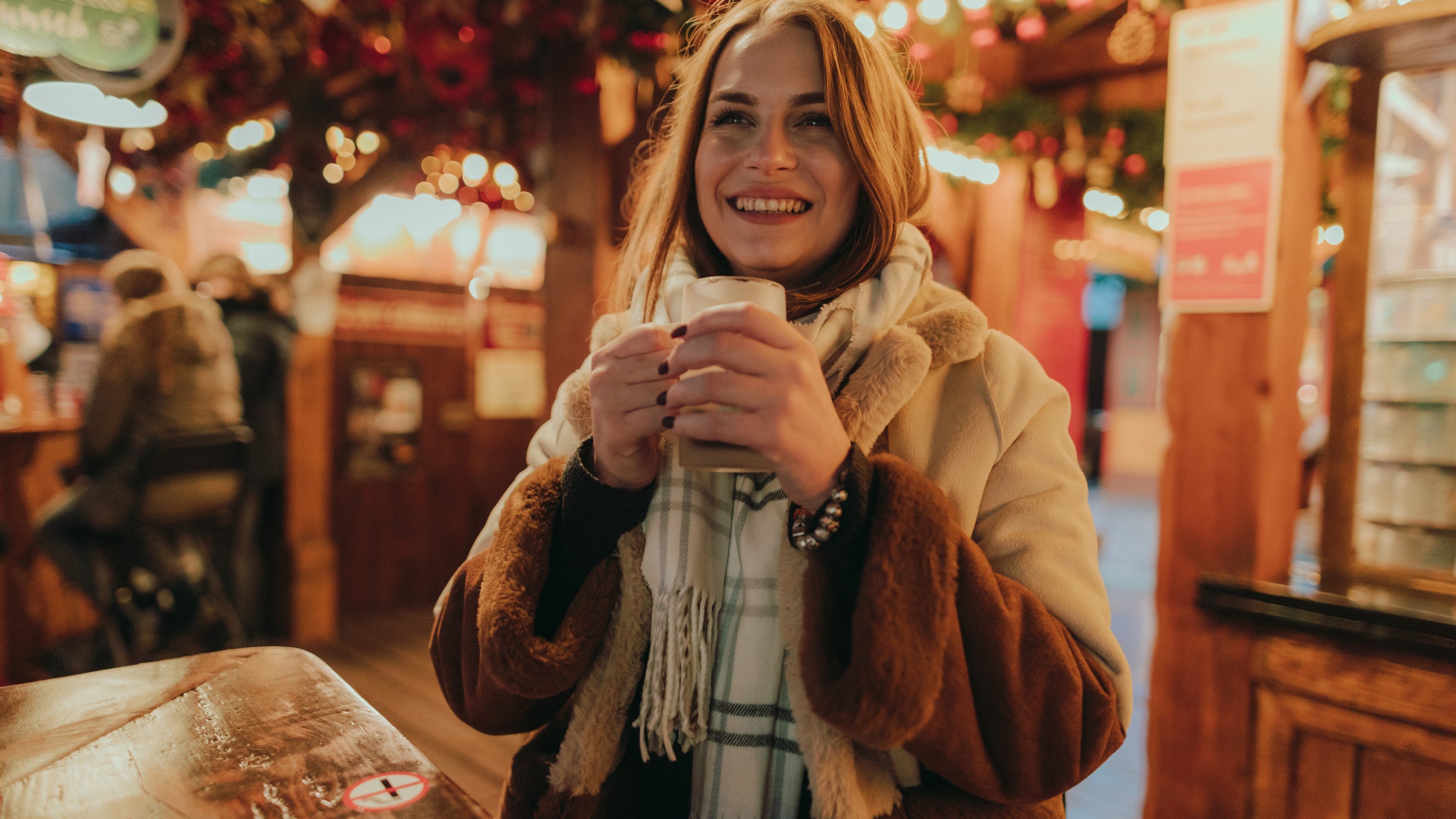 Eine junge Frau ist auf einem Weihnachtsmarkt und hält lächelnd eine Tasse Glühwein in der Hand.