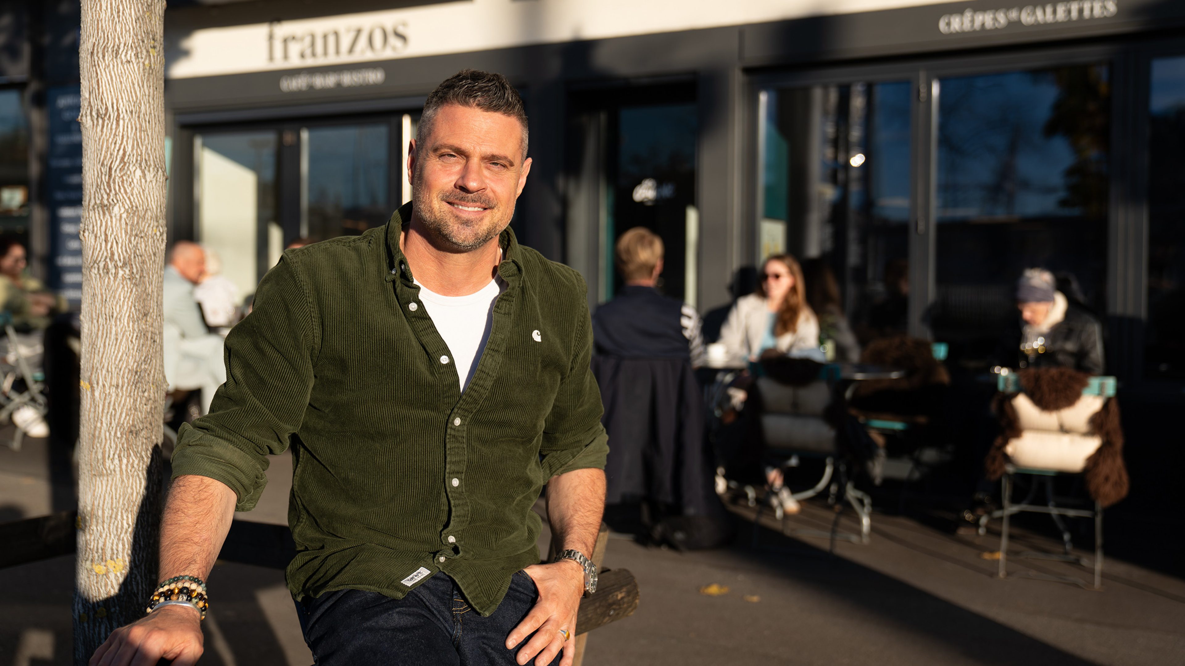 Eine Person steht mit entspannter Haltung neben einem Baum vor dem Bistro «Franzos». Sie trägt ein dunkelgrünes Hemd und eine helle Hose. Die Szene ist sonnig. Im Hintergrund sind unscharf Personen zu erkennen, die vor dem Bistro sitzen.