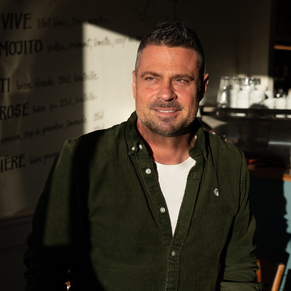 Eine Person steht in einem Bistro. Sie trägt ein dunkelgrünes Hemd und ein weisses T-Shirt darunter. Im Hintergrund ist eine helle Wand mit aufgedrucktem Text sichtbar. Rechts im Bild sind mehrere Gläser und Behälter auf einer Theke erkennbar. Das Licht fällt von links ins Bild und erzeugt deutliche Schatten.