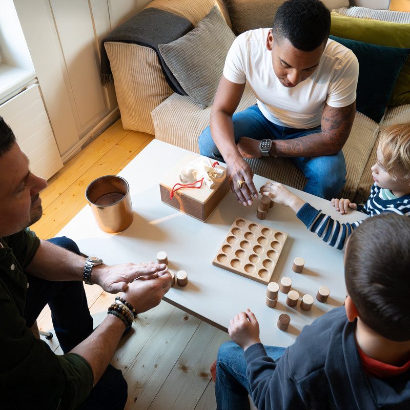 Mehrere Personen sitzen um einen niedrigen Tisch und beschäftigen sich mit einem Holzspiel. Auf dem Tisch liegen runde Holzsteine und ein Steckbrett. Der Raum hat helle Holzböden, ein Sofa im Hintergrund und neutrale Farben in der Einrichtung.
