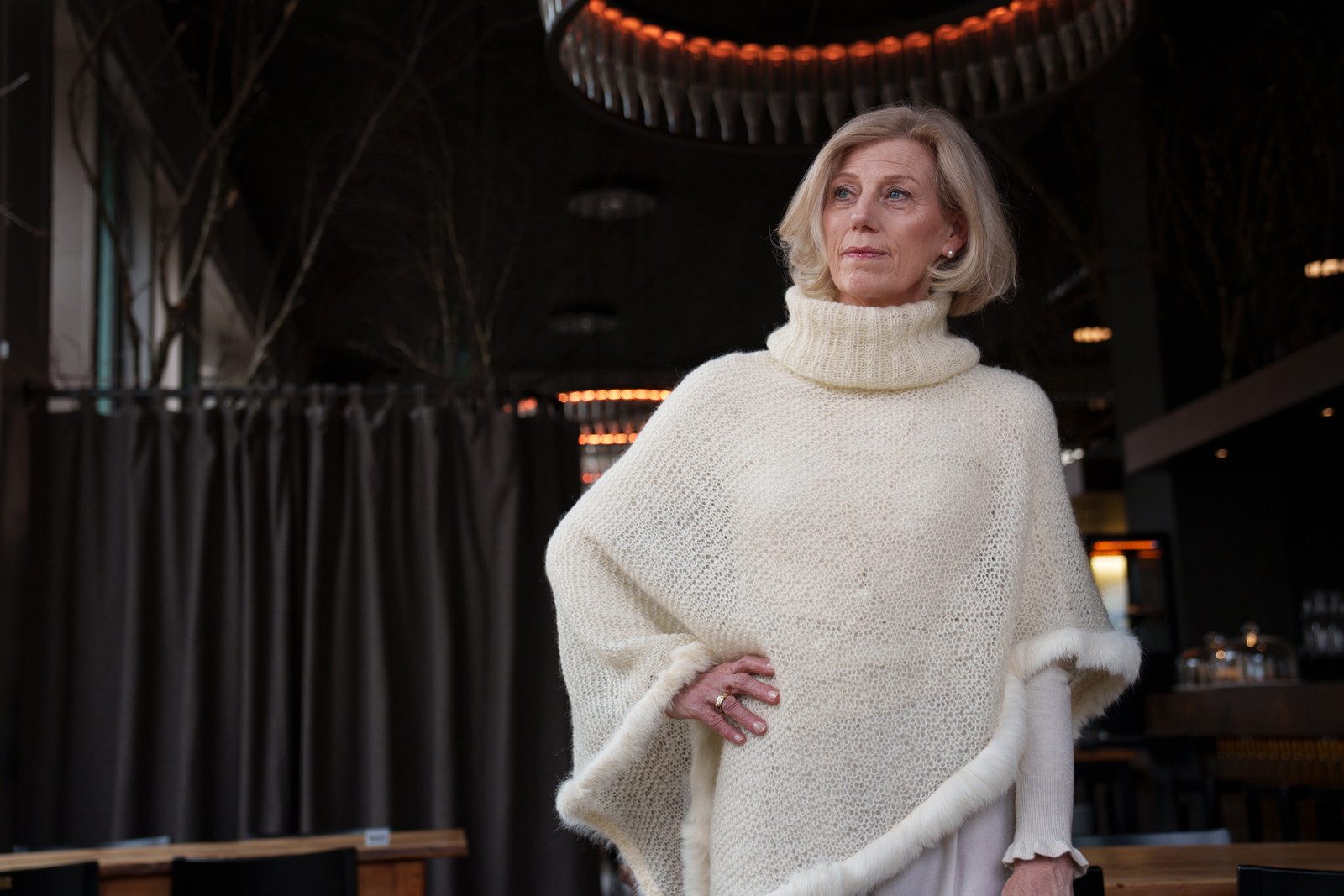 Eine ältere Frau mit einem eleganten Kurzhaarschnitt steht vor einem schwarzen Raum und blickt in eine Ecke. Sie trägt einen gestrickten, cremefarbenen Poncho und posiert mit ihrer Hand auf der Taille.