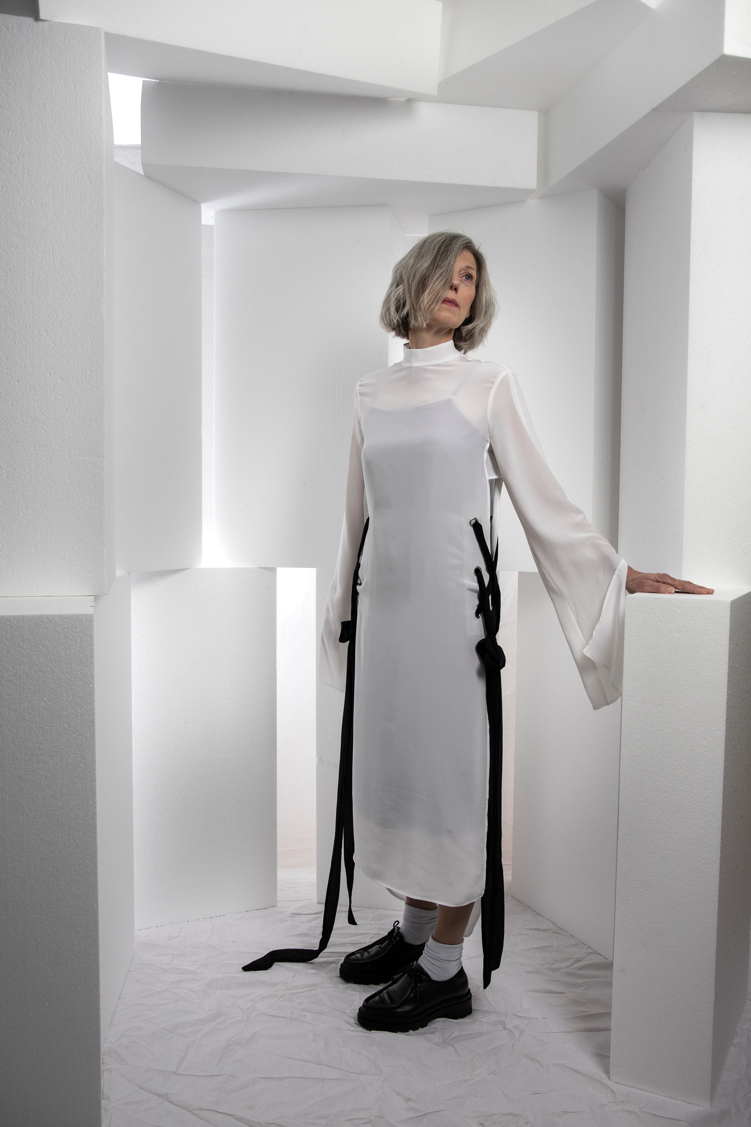 Eine mittelalte Frau in einem schwarz-weissen Kleid und mit kurzen grauen Haaren steht in einem Raum voller weisser Podeste. Sie hat die linke Hand auf einem dieser Podeste und blickt hoch, während ihre Haare ihr rechtes Auge bedecken.