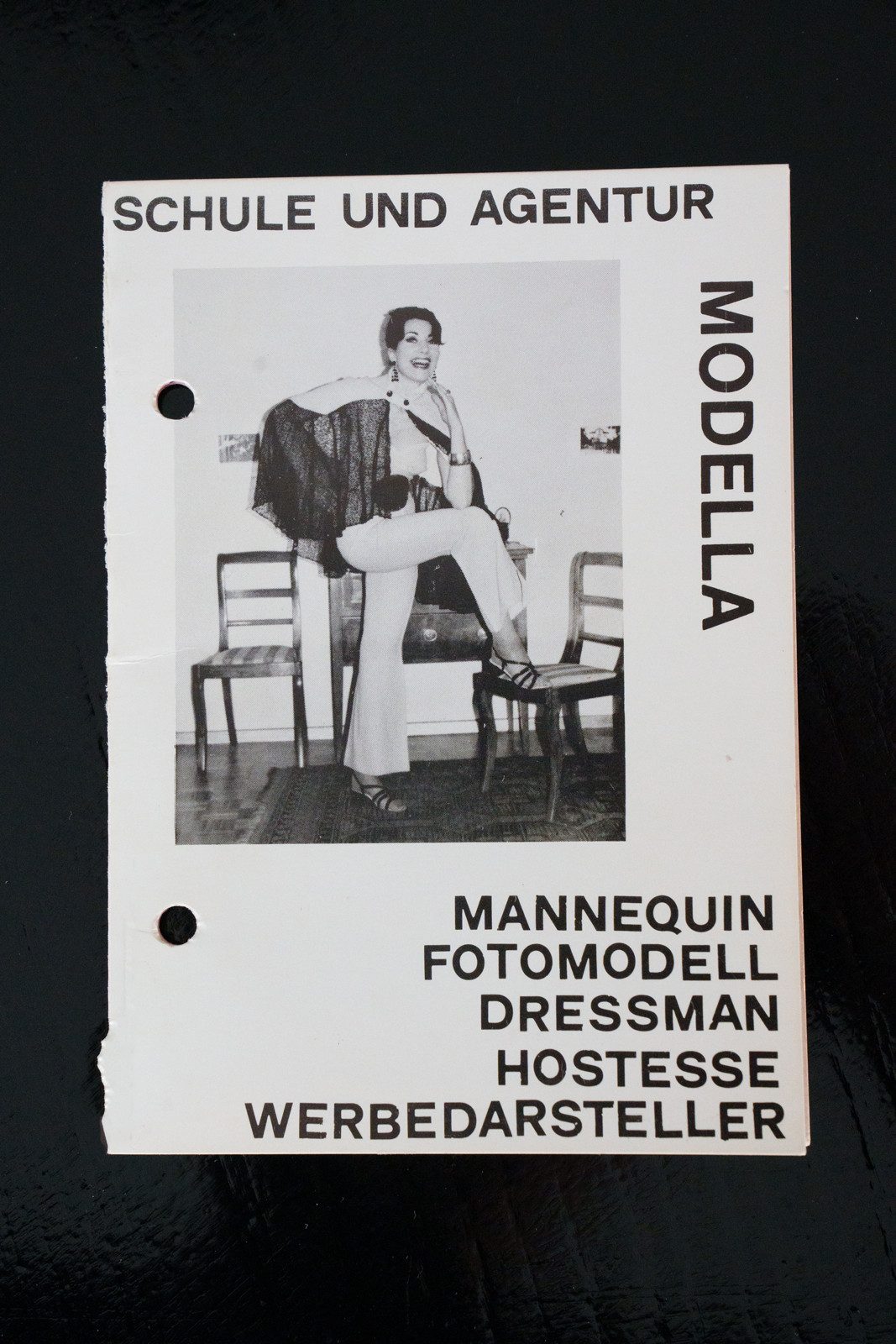 Zu sehen ist ein Flyer der Schule und Agentur Modella. Mittendrauf ist ein schwarz-weisses Foto einer jungen Frau zu sehen. Sie trägt einen weissen Jumpsuit mit exzentrischen schwarzen Elementen und schwarze Sandalen. Unter dem Bild sind einzelne Ausbildungsbereiche aufgezählt: Mannequin, Fotomodell, Dressman, Hostesse und Werbedarsteller.