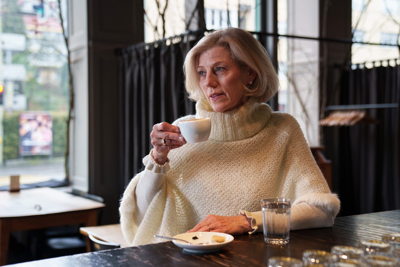 Eine ältere Frau mit einem eleganten Kurzhaarschnitt und einem cremefarbenen Poncho sitzt an einem schwarzen Tisch und hält eine weisse Kaffeetasse in der Hand. Neben ihr stehen ein Glas mit Wasser und ein Untersatzteller mit Keks zum Kaffee.