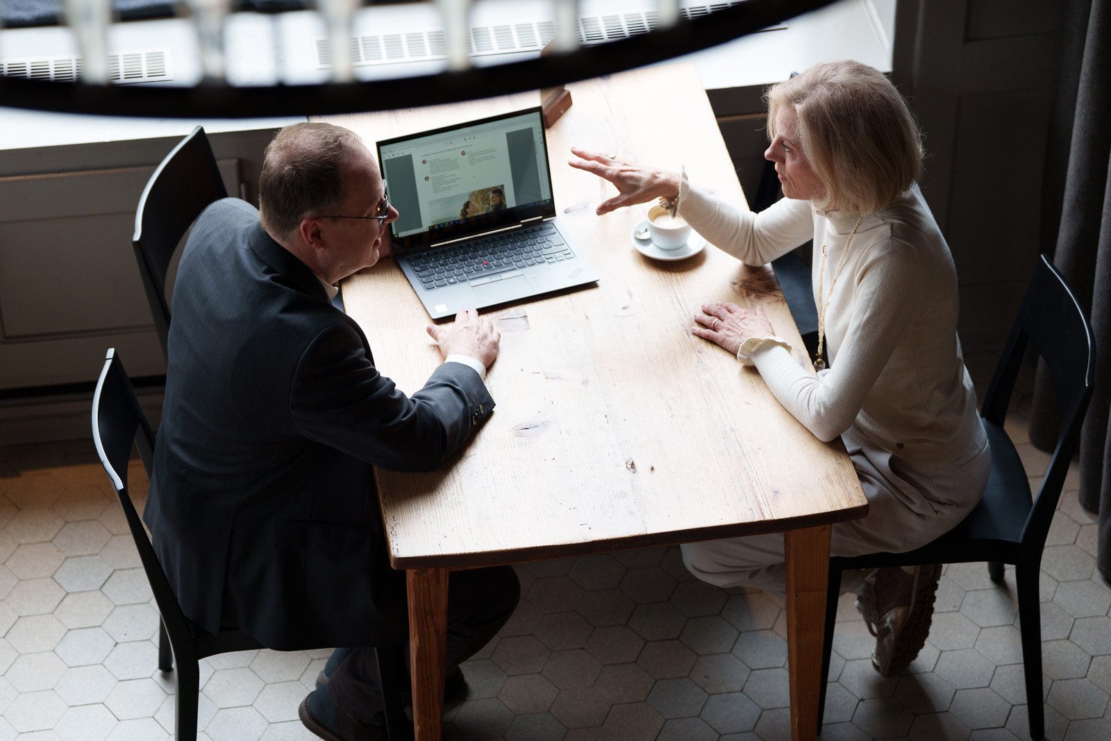 Ein Mann mit Brille und schwarzem Blazer führt ein Gespräch mit einer kurzhaarigen, hell gekleideten Frau. Neben ihnen auf dem Tisch stehen ein Laptop und eine Kaffeetasse.