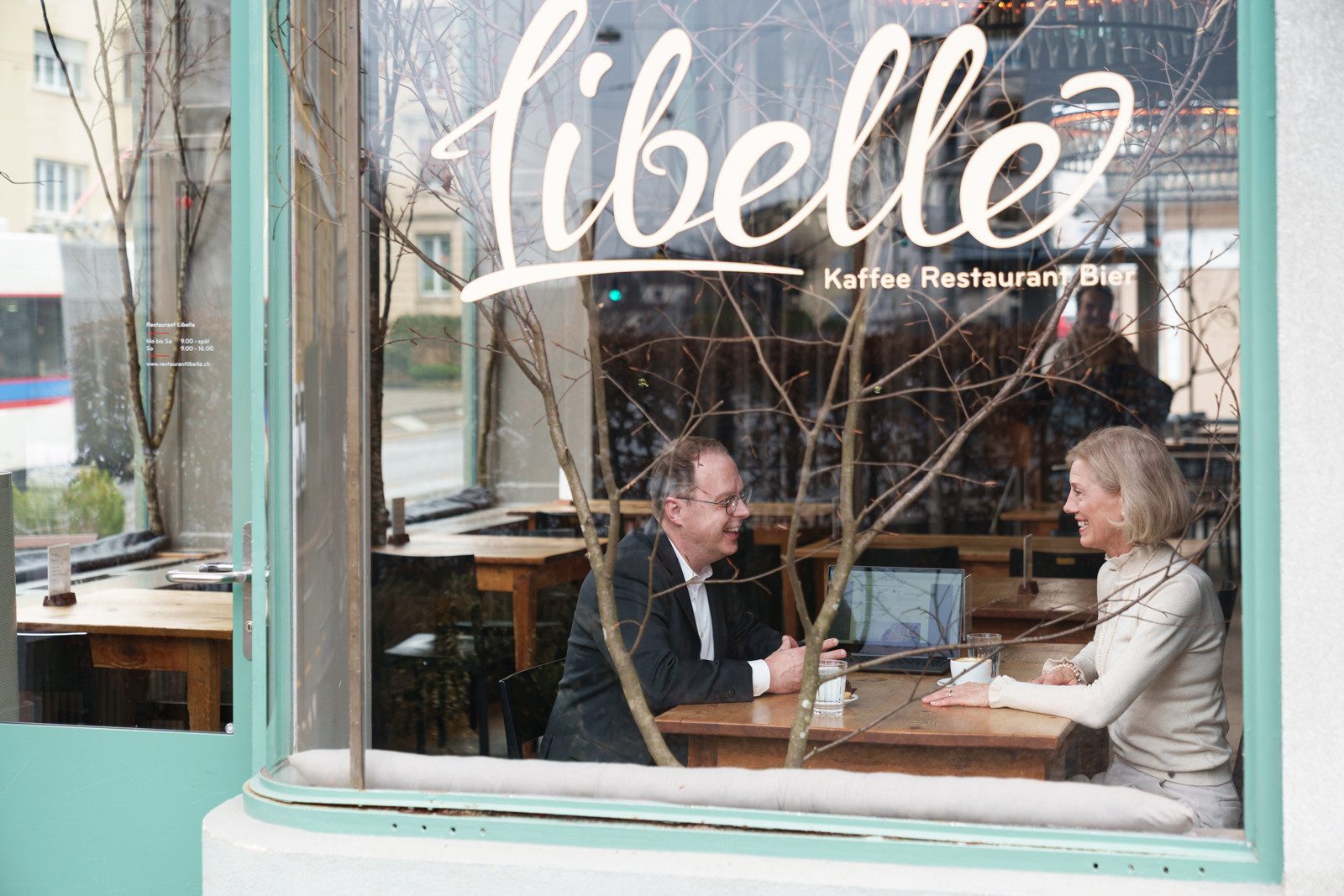 Durch ein grosses Fenster mit der Beschriftung «Libelle – Kaffee, Restaurant, Bar» blickend, sieht man zwei lachende Personen an einem Holztisch im Gespräch mit Kaffee. Der Mann trägt einen schwarzen Blazer mit weissem Hemd und die Frau ist cremeweiss gekleidet. Zwischen ihnen steht ein offener Laptop.