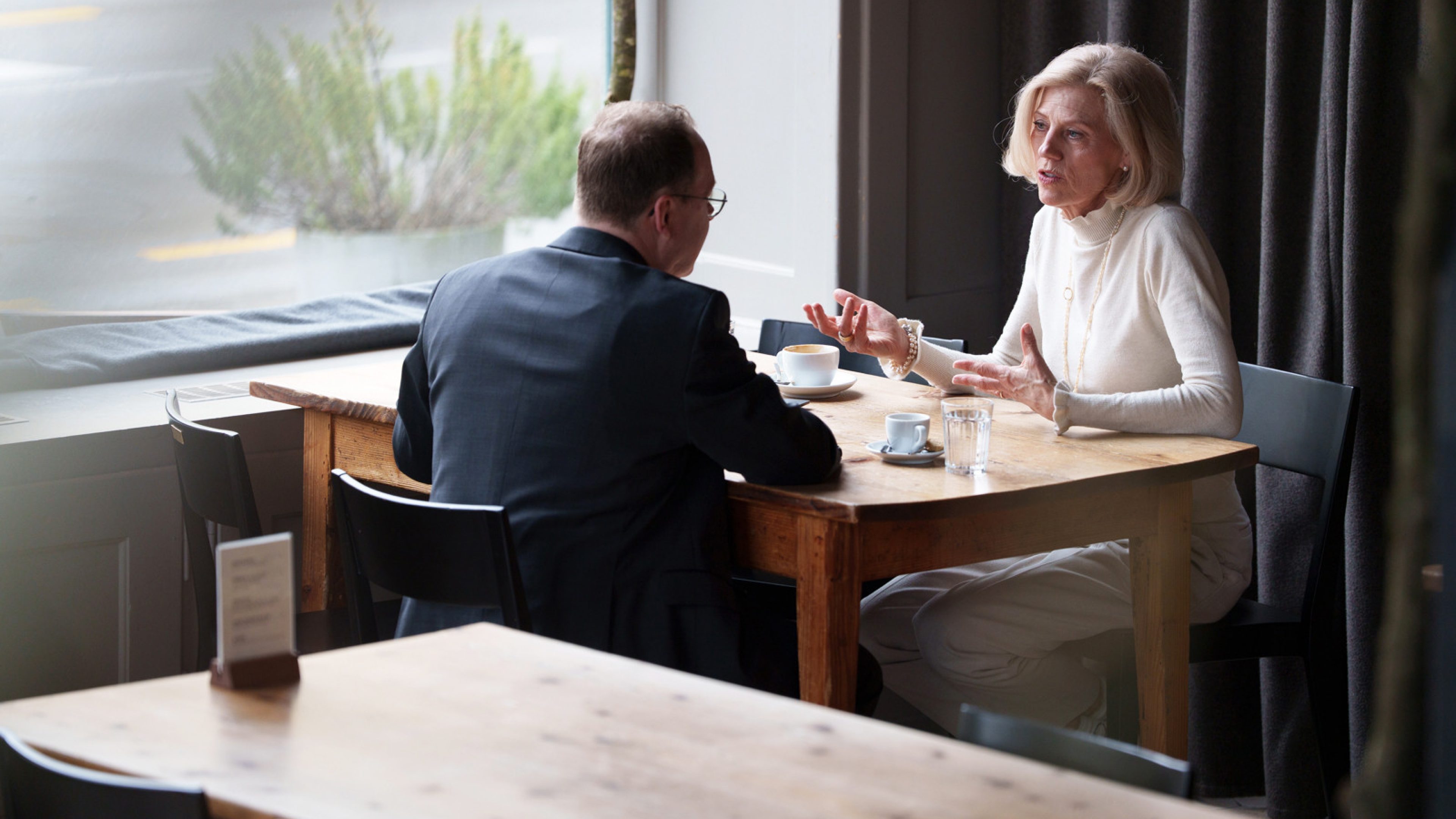 Ein Mann mit Brille und schwarzem Blazer führt ein Gespräch mit einer kurzhaarigen, hell gekleideten Frau. Neben ihnen auf dem Tisch stehen ein Laptop und eine Kaffeetasse.