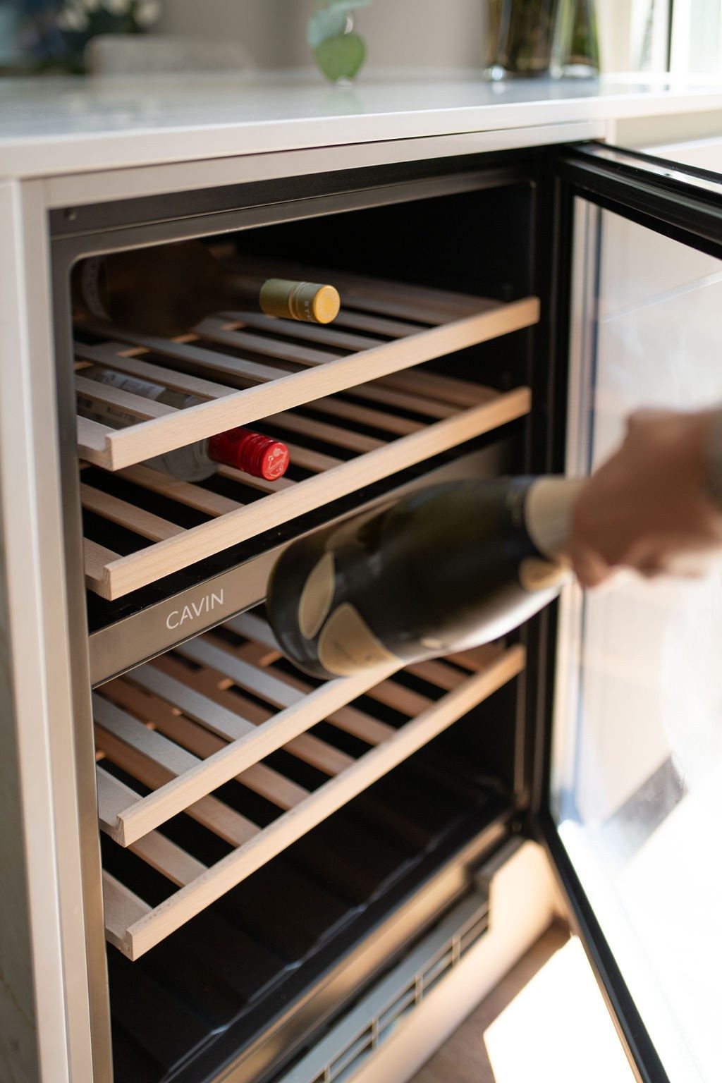 Eine Flasche wird aus einem Weinkühlschrank genommen. Der Weinkühlschrank hat eine Glastüre und innen verschiedene Holzregale, auf deinen der Wein gelagert werden kann.
