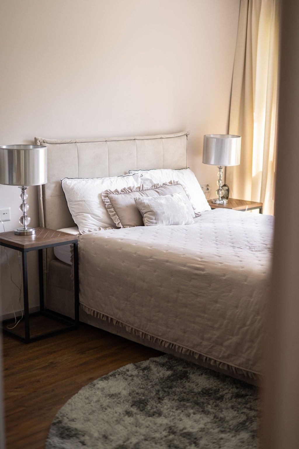 Ein Ausschnitt eines Schlafzimmers in grauen Tönen mit Hotel-Flair. Auf dem Bett sind vier Kissen angeordnet und eine grosszügige Tagesdecke. Neben dem Bett stehen zwei Nachttischen mit identischen Nachttischlampen.