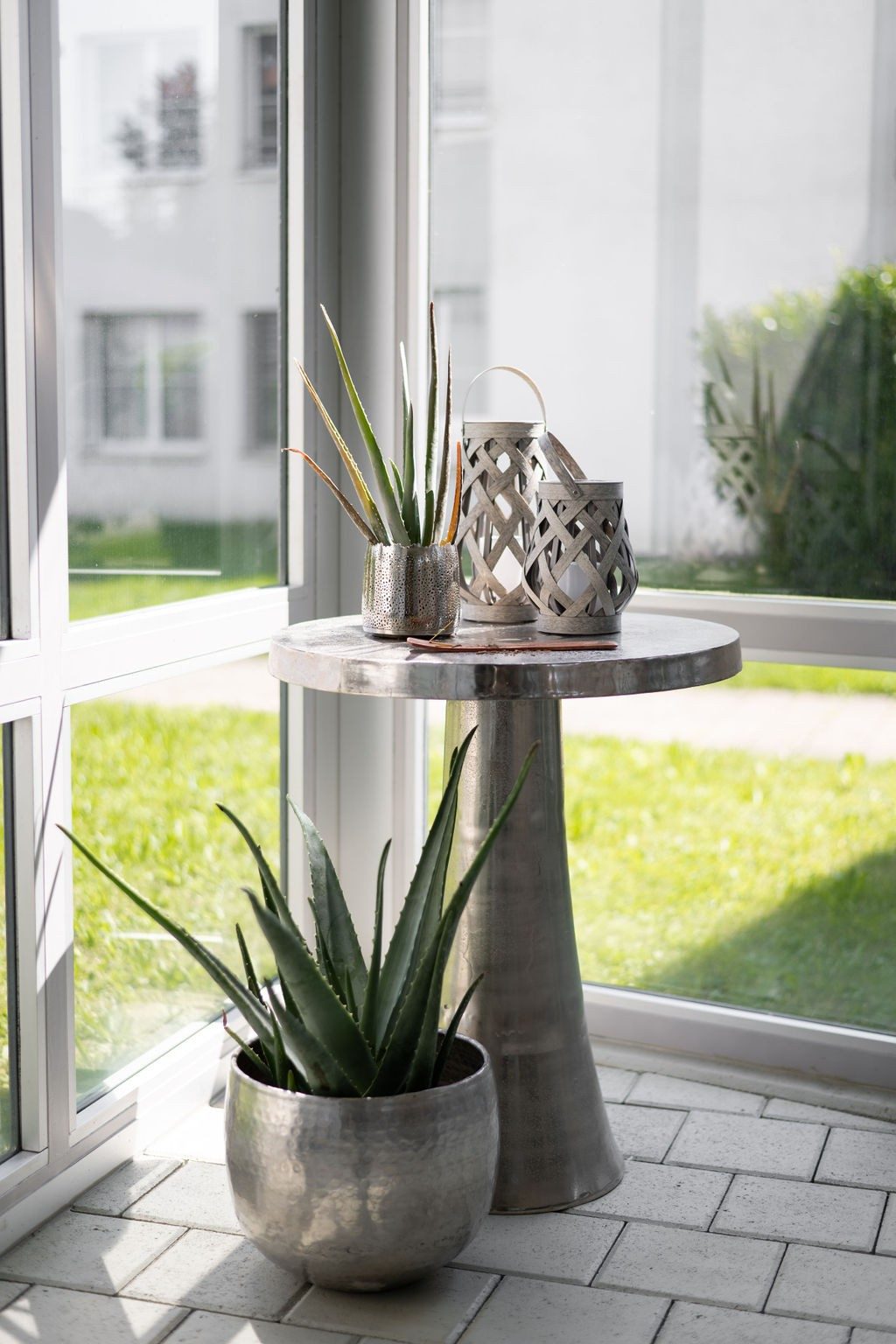 Ein runder metallischer Tisch steht in der Ecke eines Wintergartens. Auf und neben dem Tisch sind Aloe-Vera -Pflanzen in Töpfen im Metallic-Look.