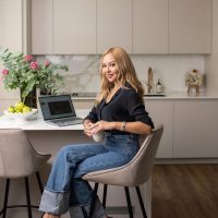 Eine blonde Frau sitzt auf einem Barhocker an einer Kochinsel und blickt über ihre Schulter lächelnd in die Kamera. Sie macht etwas an einem Laptop, neben dem ein prächtiger Blumenstrauss steht.