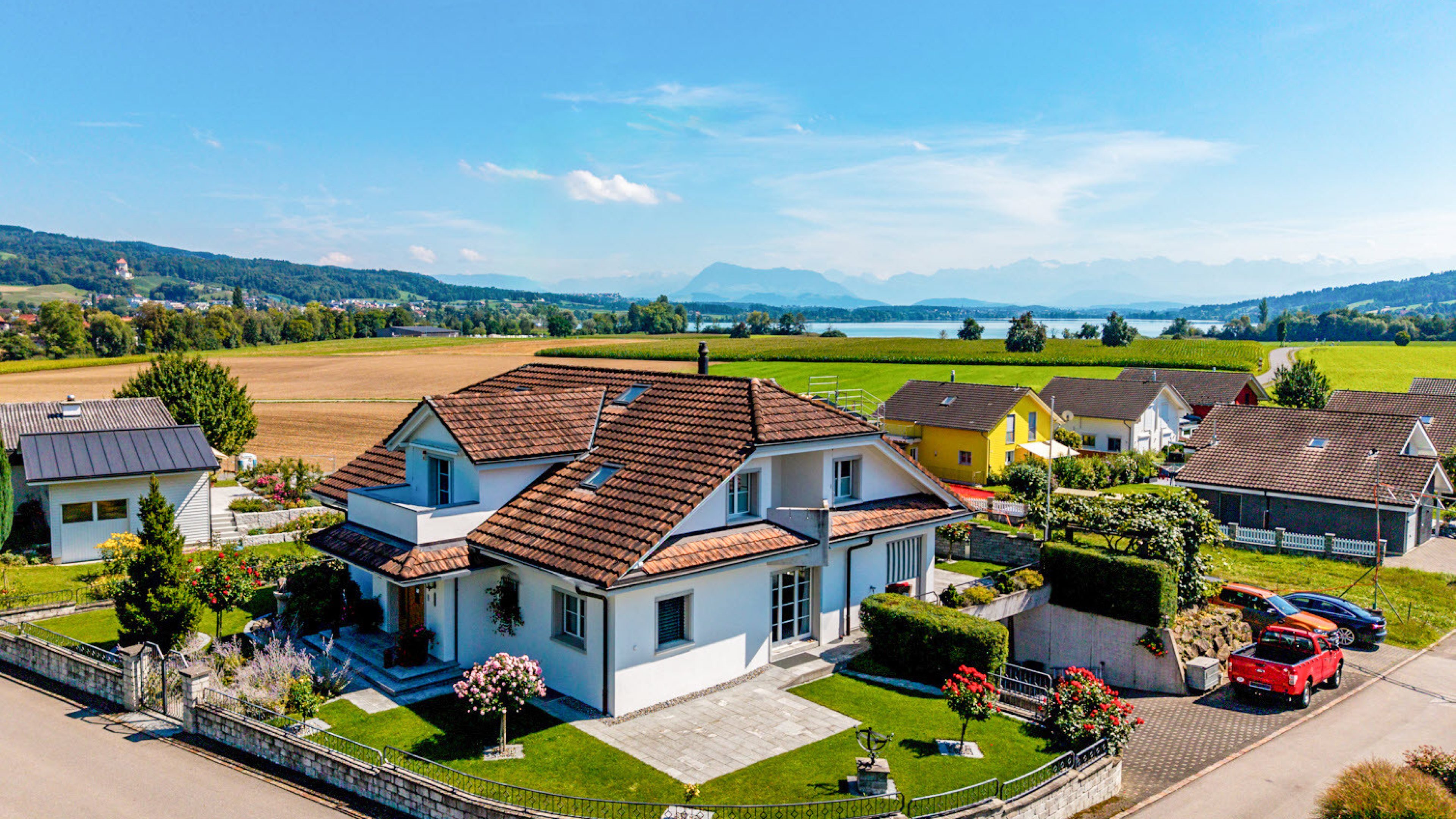 Freundliches, weisses Einfamilienhaus mit braunem Ziegeldach, umgeben von gepflegter Grünfläche. Es gibt noch ein paar Nachbarshäuser und im Hintergrund sieht man einen See und Berge.
