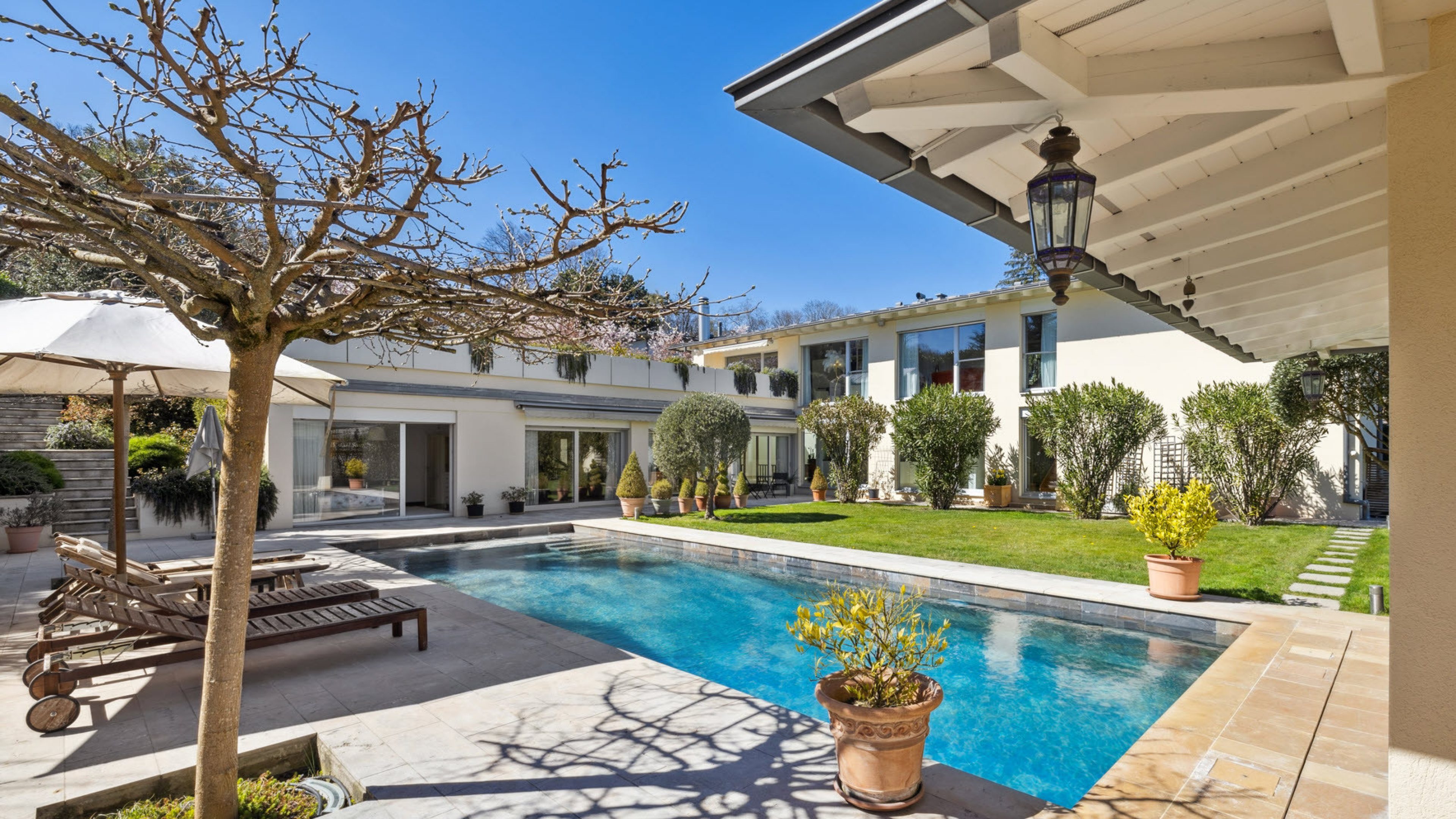 Ein weisses Haus mit Garten und grossem Pool. Um den Pool herum hat es verschiedene Pflanzen, Liegebetten und Sonnenschirme. Der Himmel ist blau.