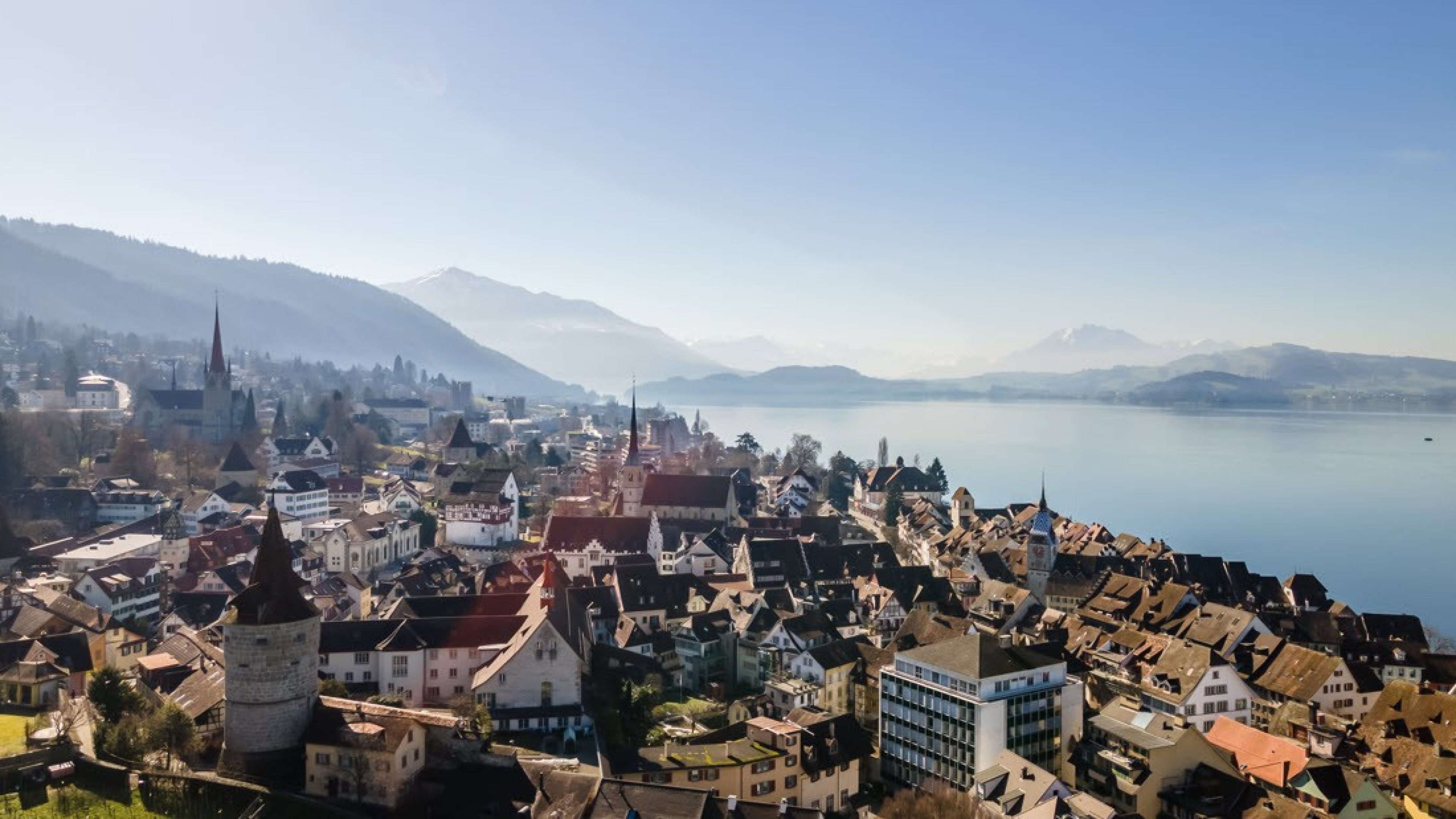 Luftaufnahme von der Stadt Zug und dem Zugersee. Im Hintergrund die Bergkulisse im Herbstnebel.