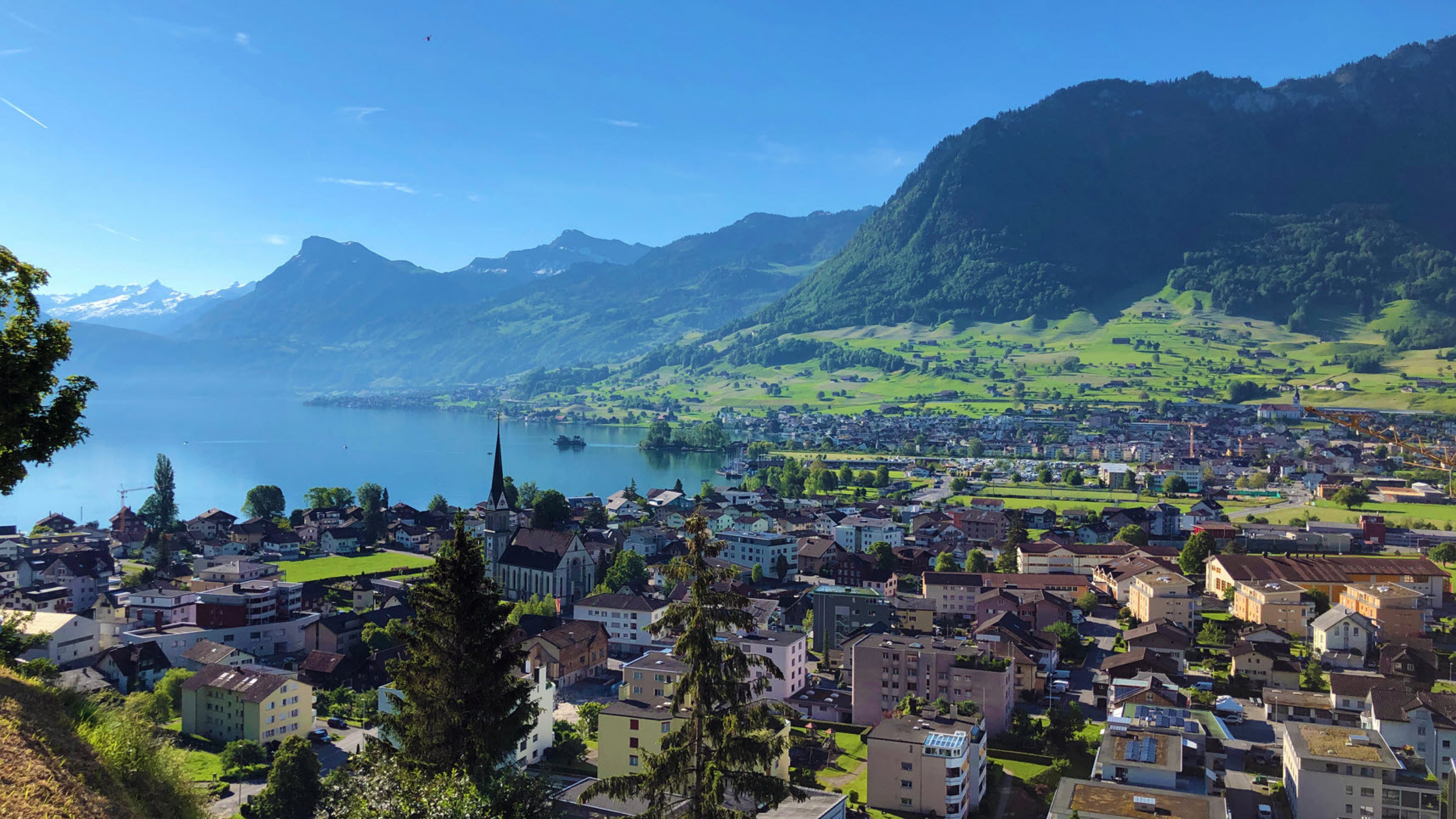 Sicht auf Siedlung Ennetbürgen am Ufer des Luzerner Sees und am Ende des Engelbergertals im Kanton Uri. Die Felder und die Wälder sind grün, der Himmel ist blau.