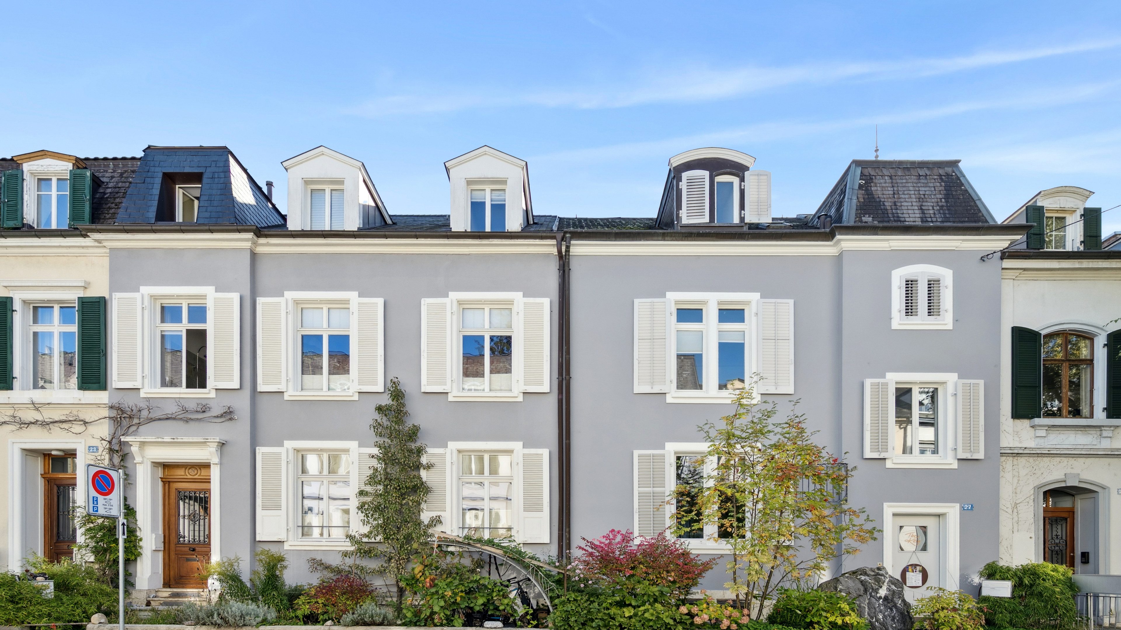 Zweistöckige helle Reihenhäuser mit weissen oder grünen Fensterläden. Der Himmel ist blau und vor den Häusern hat es verschiedene Sträucher.