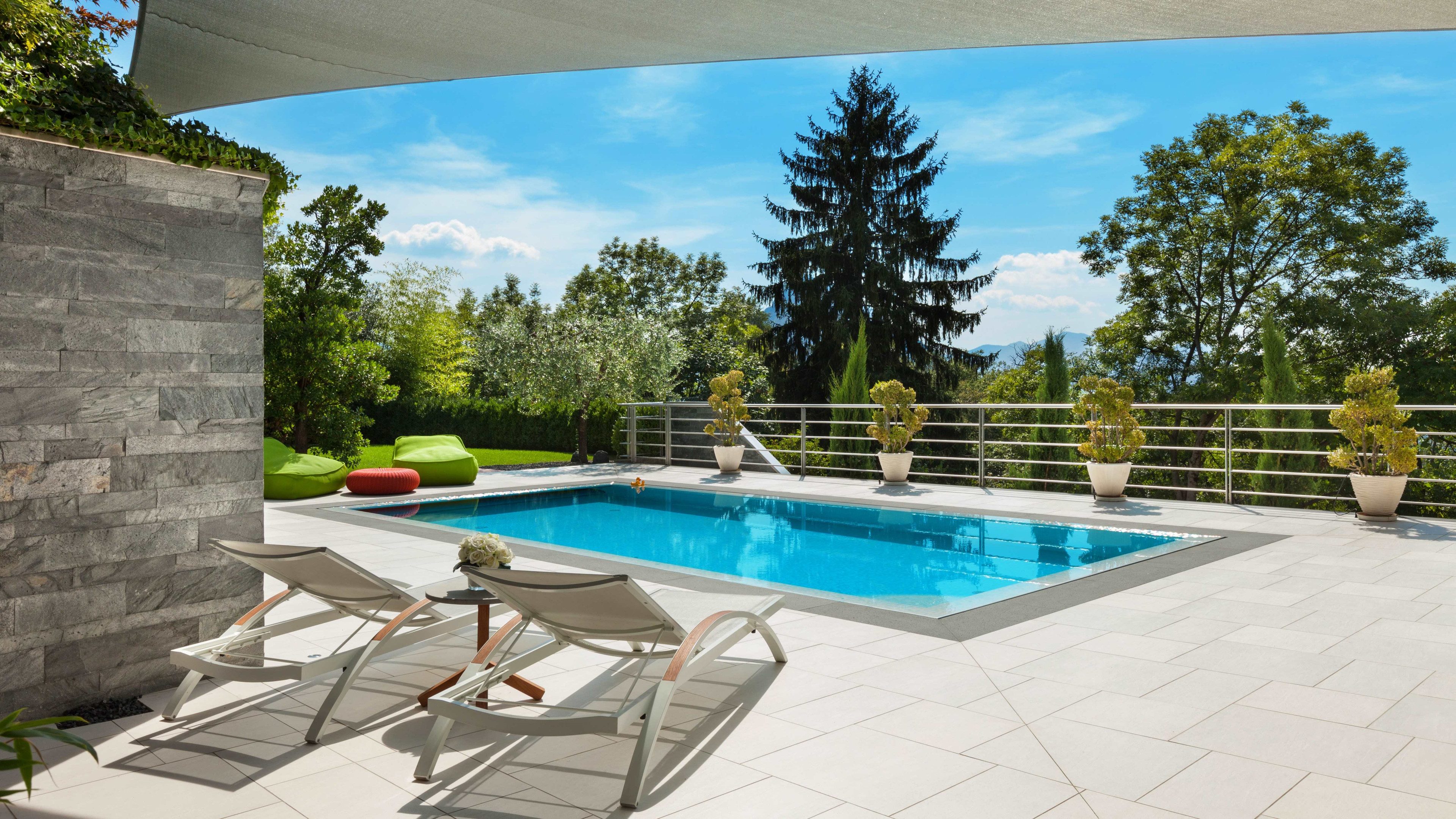 Hellblauer Pool umgeben von beigen Platten. Zwei Liegestühle und zwei Stoffsessel, sowie diverse Topfplanzen befinden sich auf den Platten. Im Hintergrund sind Bäume, der Himmel ist blau. 
