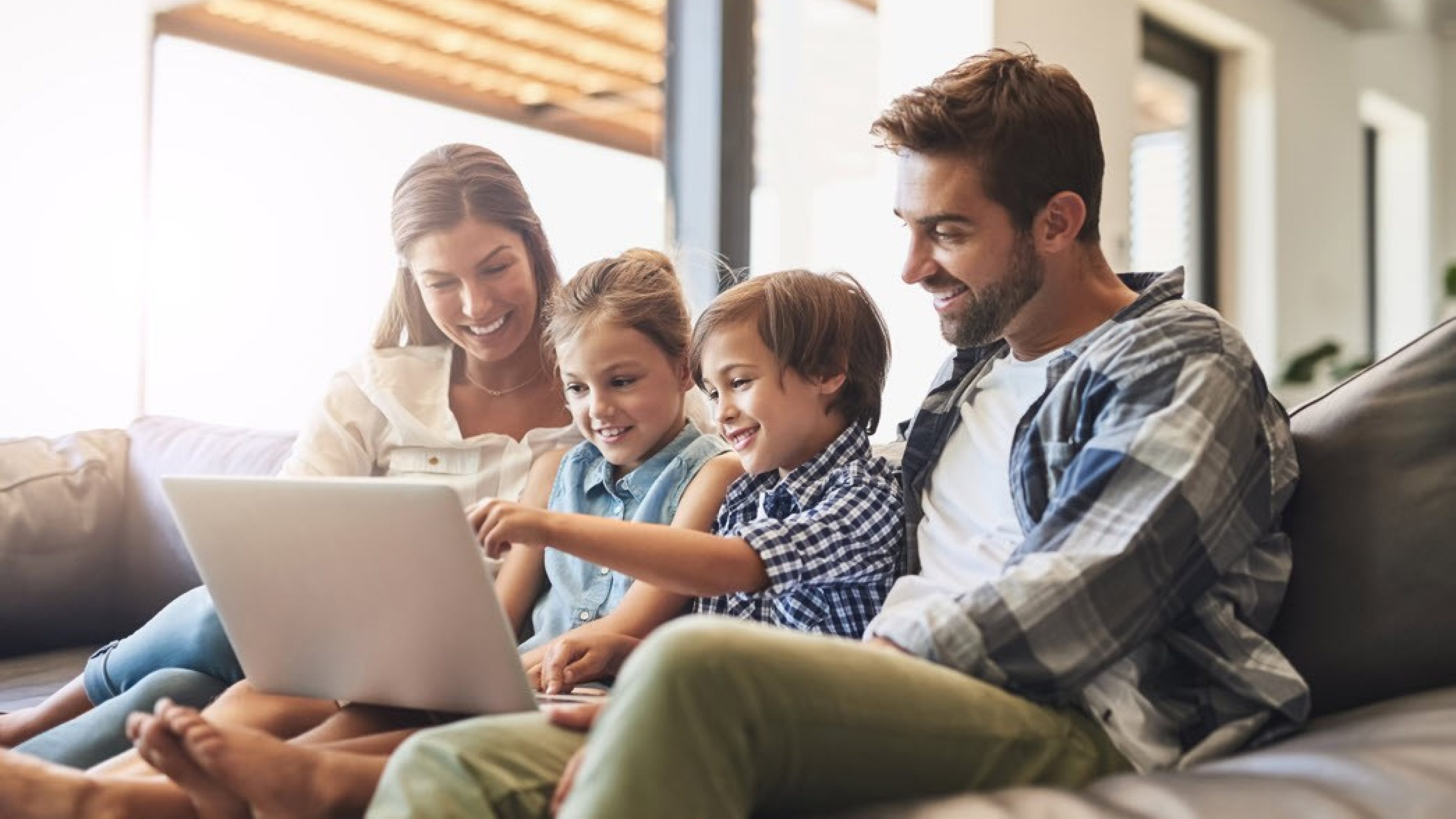 Vater, Mutter und zwei Kinder sitzen auf dem Sofa und schauen glücklich in einen Laptop.