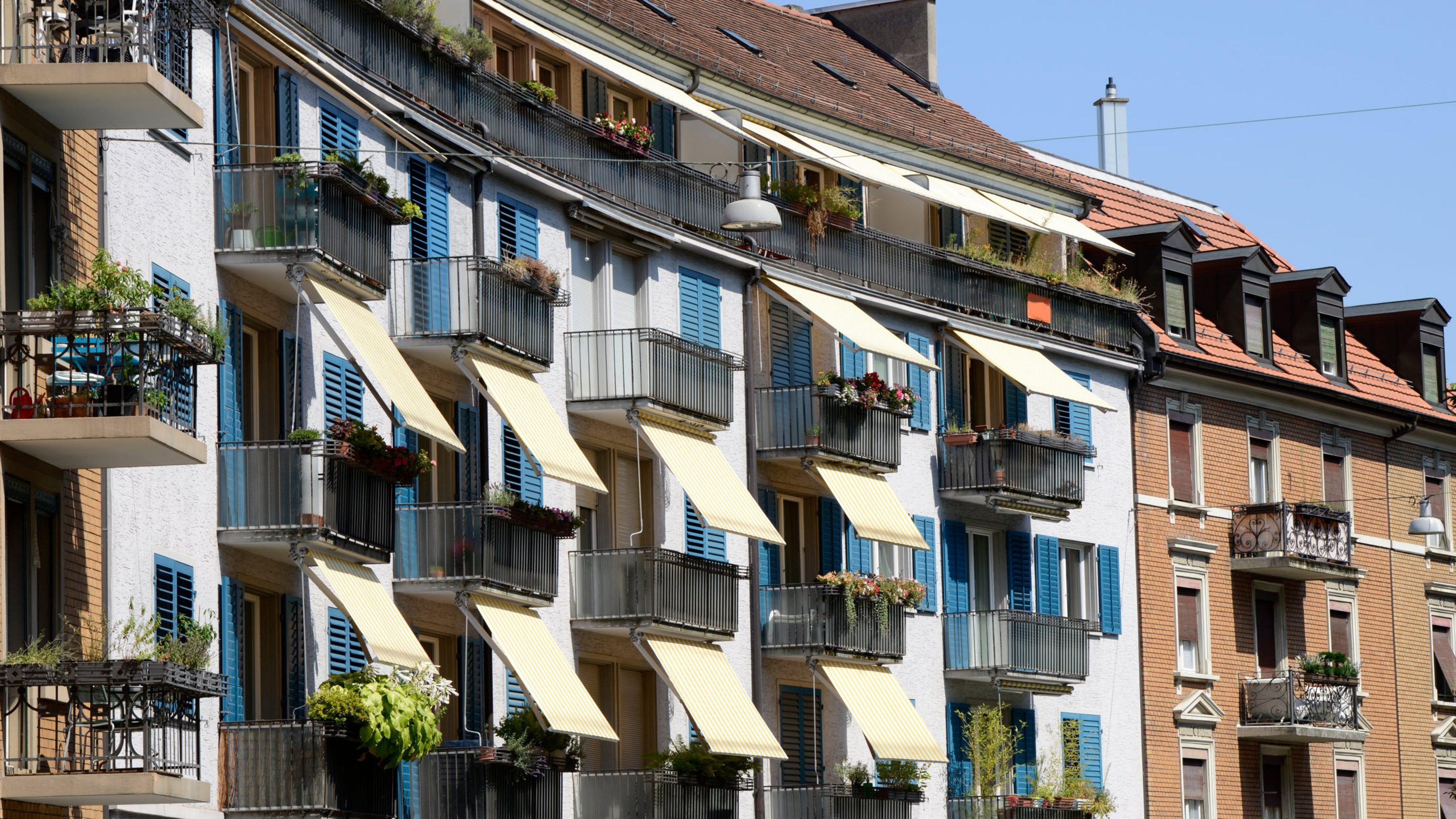 Zwischen zwei rotbraunen Hausfassaden gibt es eine weisse Fassade mit hellblauen Fensterläden. Es hat viele kleine Balkone mit Pflanzen und bei einigen sind die gelben Sonnenstoren hinausgelassen.