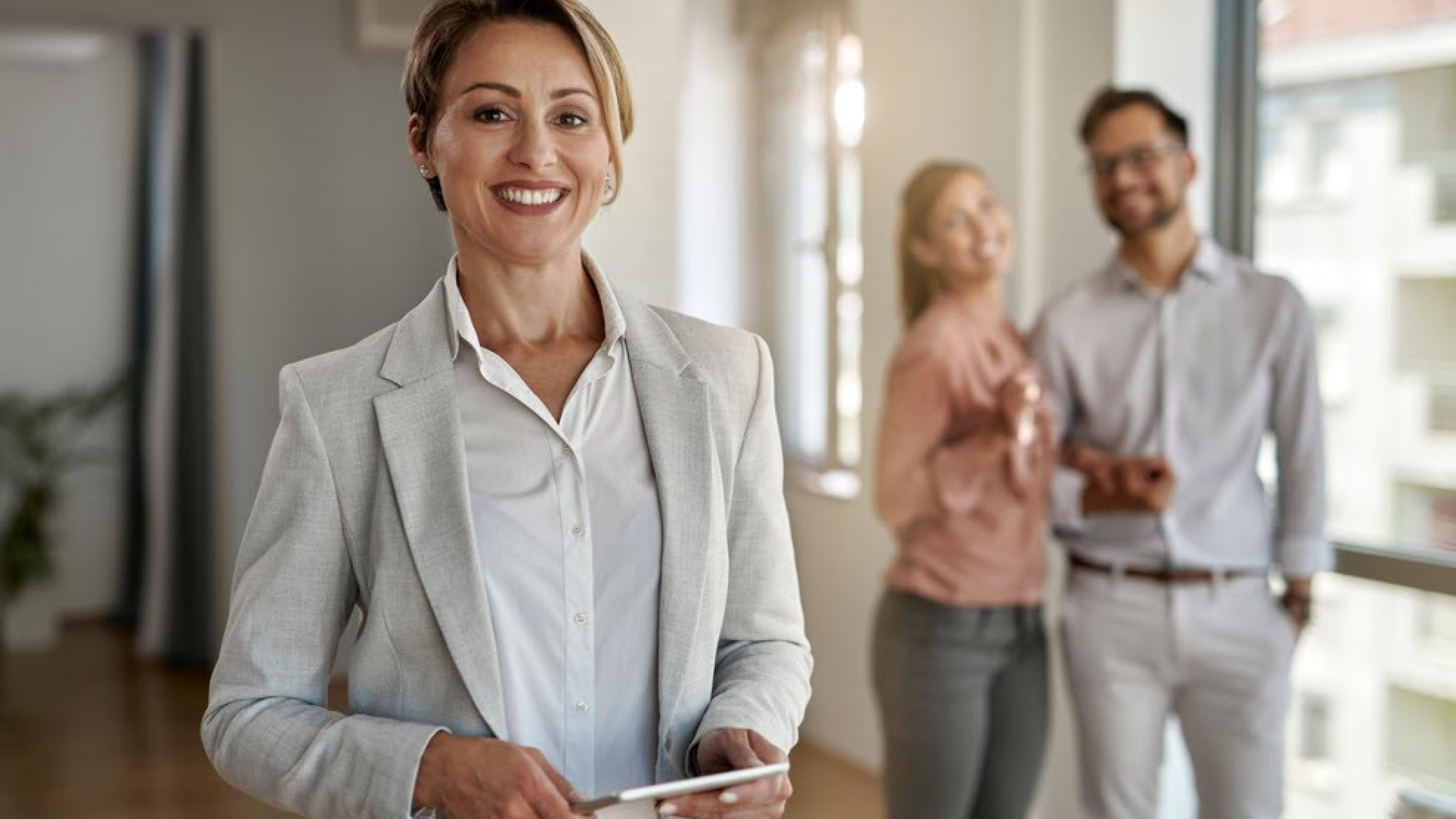 Eine Immobilienberaterin mit blonden, kurzen Haaren steht im Vordergrund. Hinter ihr befindet sich ein glückliches, junges Ehepaar.  Die Frau hält einen Schlüssel in der Hand. Alle Personen im Bild lachen.