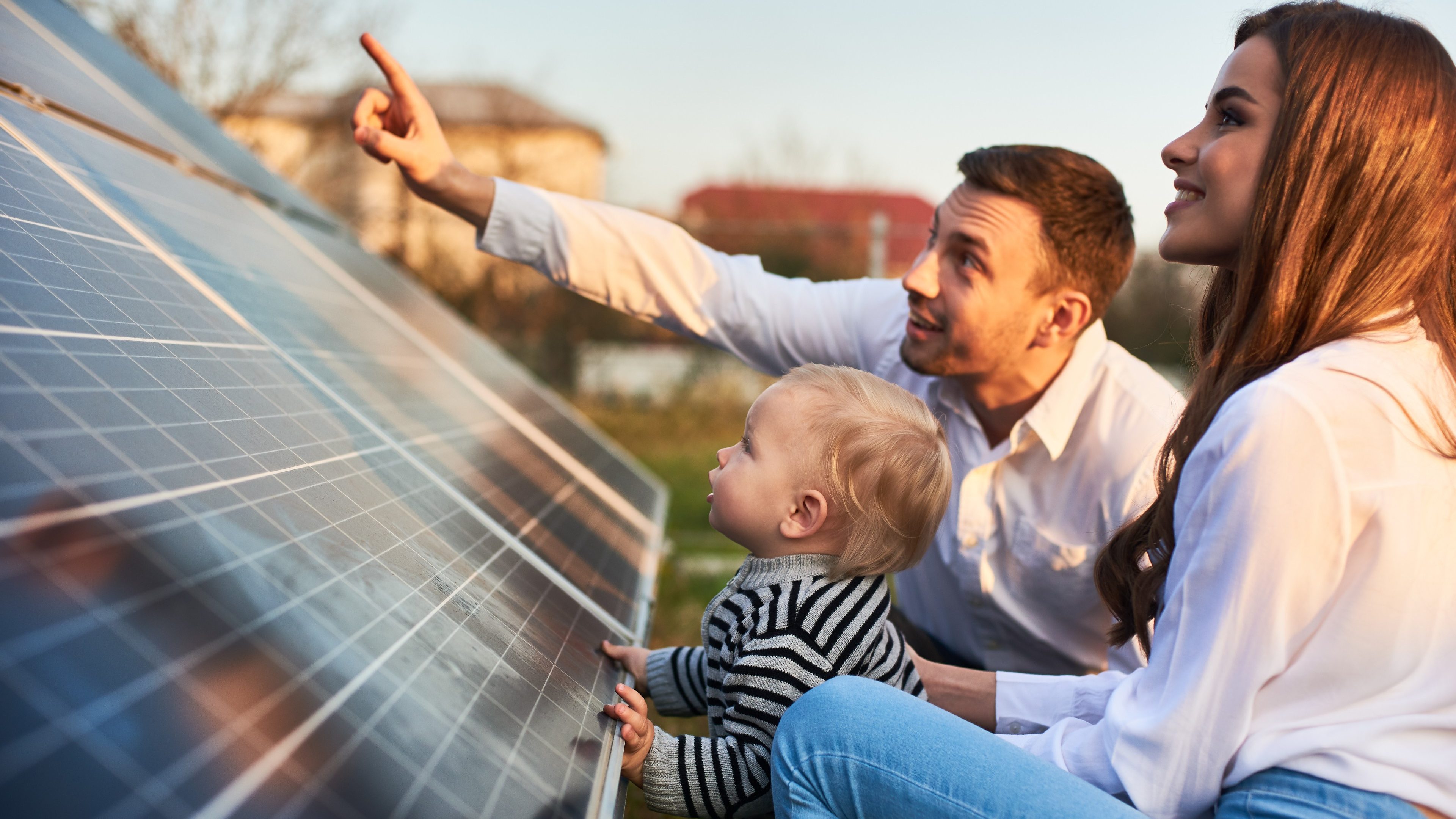 Man shows his family the solar panels on the plot near the house during a warm day. Young woman with a kid and a man in the sun rays look at the solar panels.