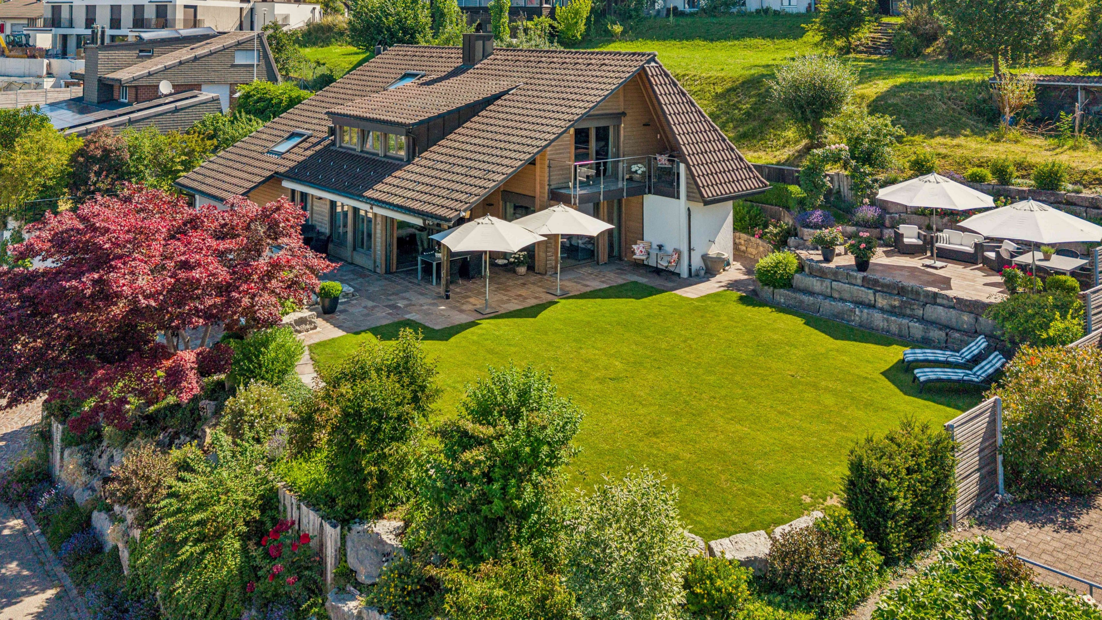 Luftansicht: freundliches Einfamilienhaus umgeben von einer grossen Grünfläche und Bäume und Sträucher. Rechts vom Haus hat es einen separaten Sitzplatz mit Möbel und zwei Sonnenschirme.