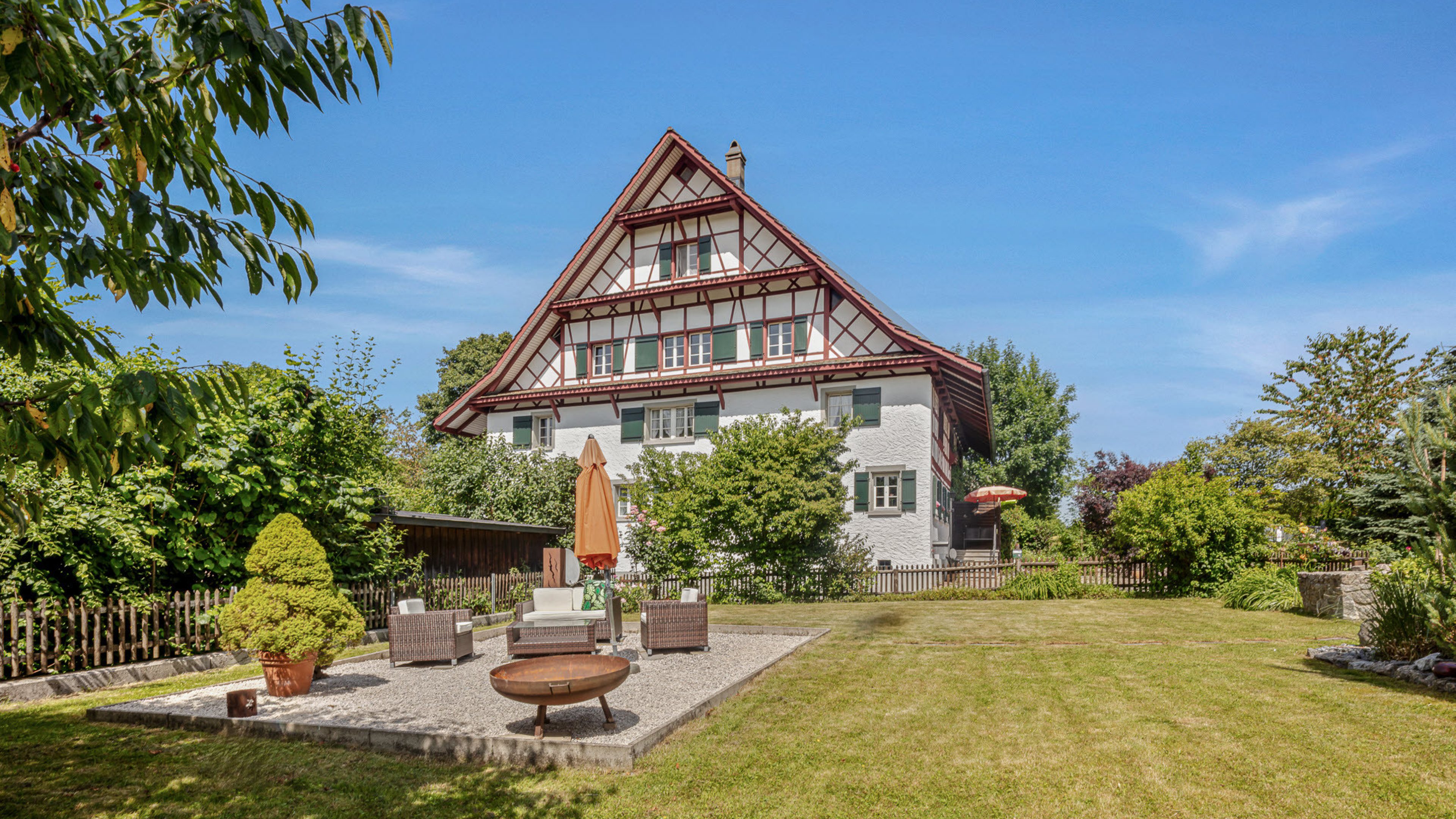 Grosses, freundliches Haus mit grosser, frisch gemähter Rasenfläche und einem Sitzplatz mit Gartenmöbel, Sonnenschirm und Grillschale. Der Himmel ist blau, die Sonne scheint.