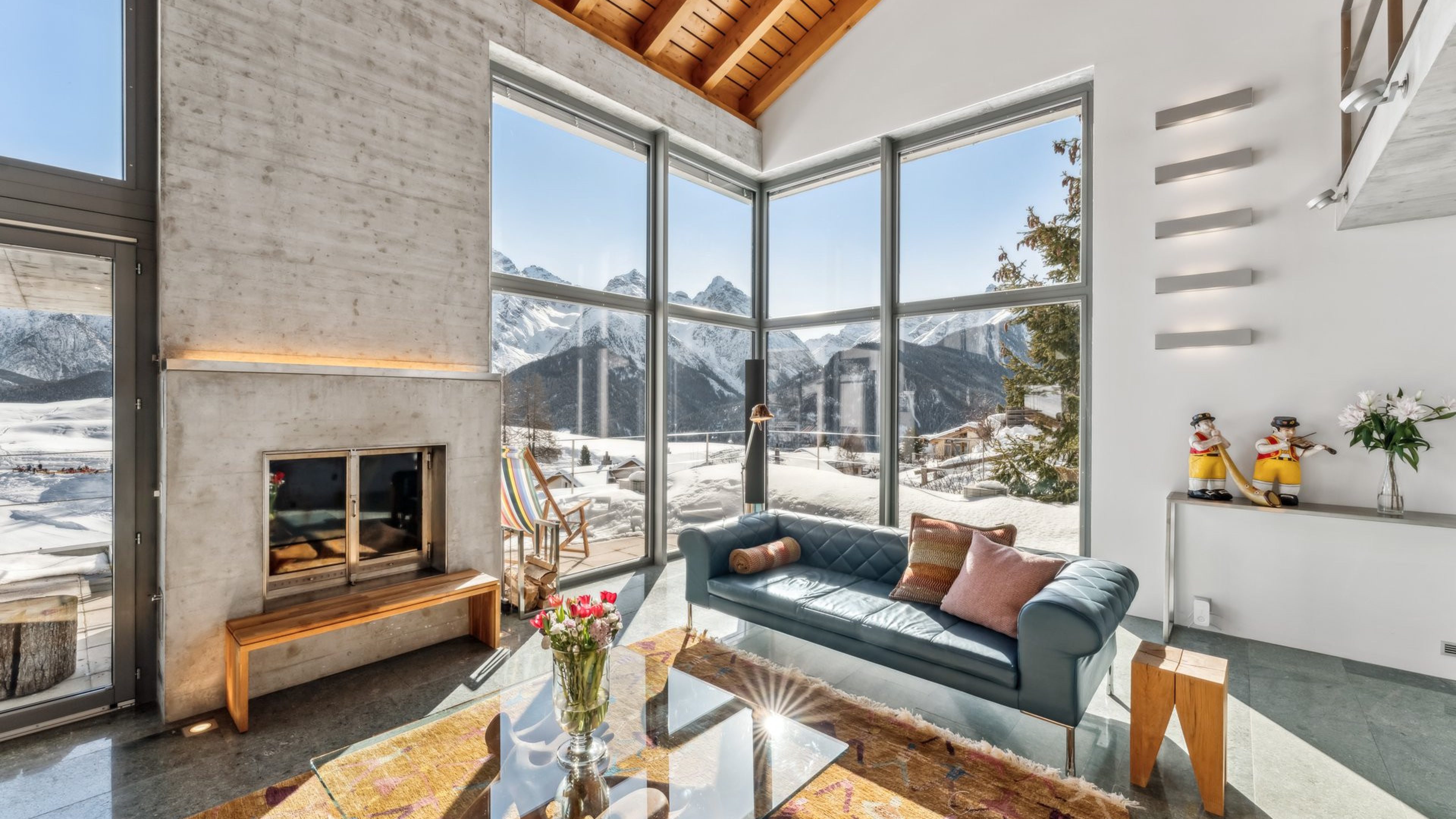 Helles Wohnzimmer mit grossen Glasfenstern, durch welche eine Schneelandschaft und Schneeberge zu sehen sind. Im Wohnzimmer befindet sich ein blaues Sofa, davor ein Teppich und ein Glastisch, auf welchem eine Vase mit Blumen steht.