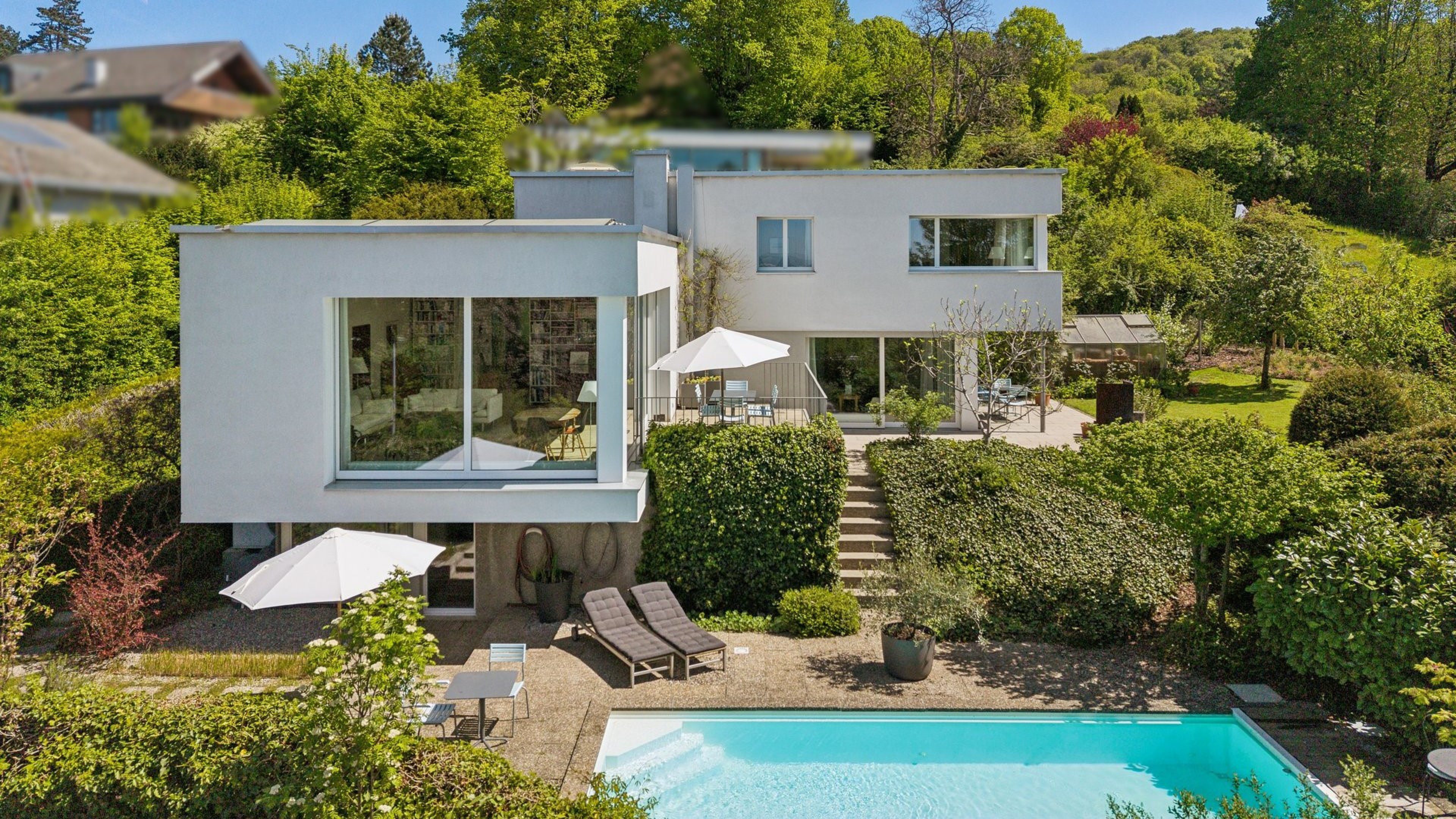 Modernes, weisses Einfamilienhaus mit Pool, umgeben von vielen Bäumen und Sträuchern. 