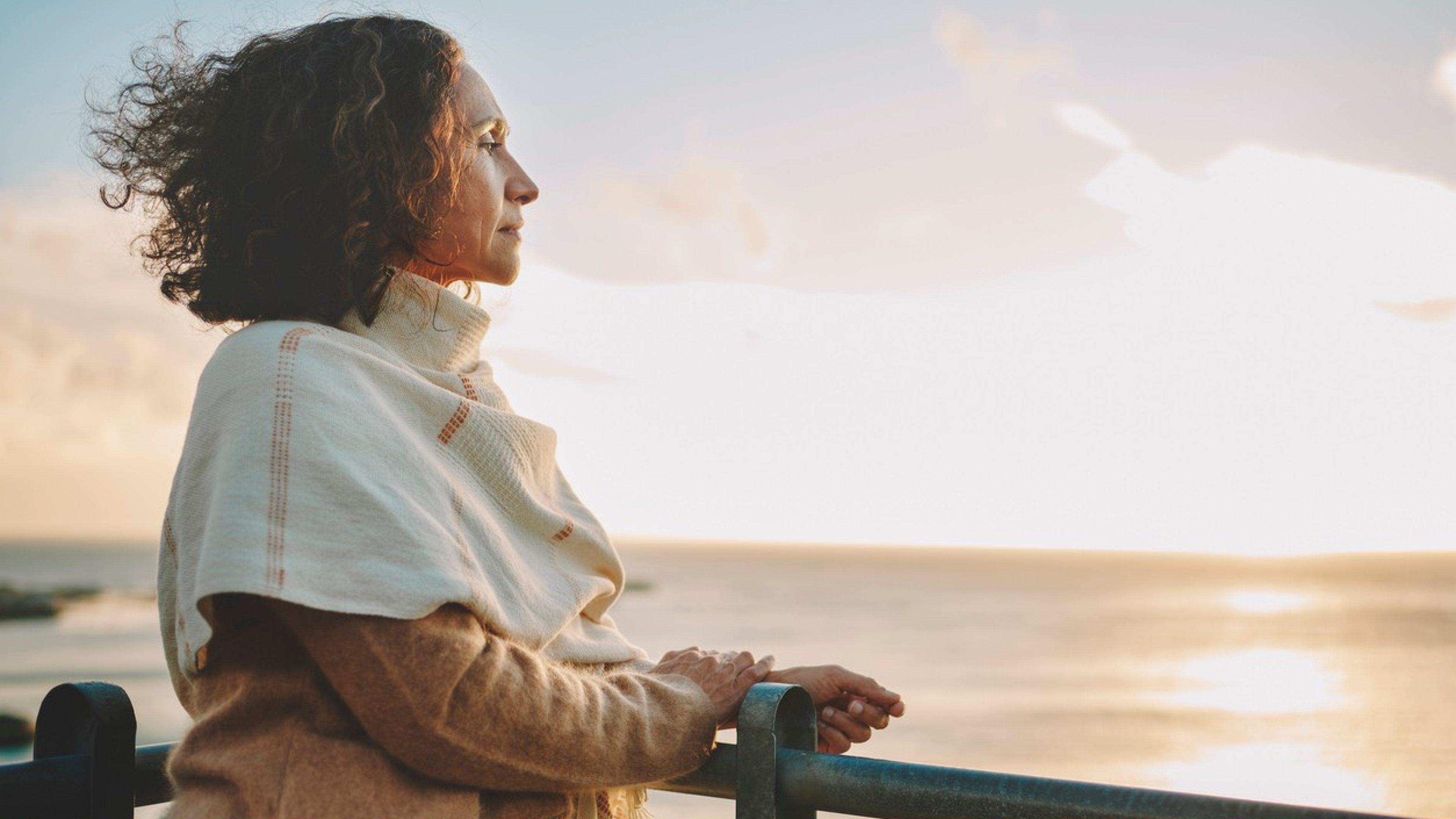 Mature woman wearing a pashmina leaning on a railing and looking out at the sunset over the ocean