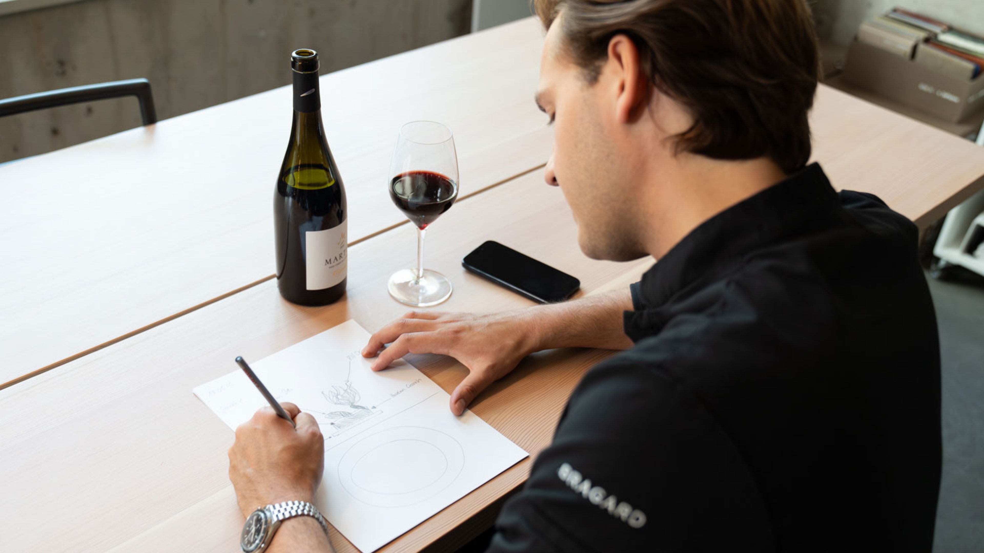 Ein junger Mann in schwarzem Kochkittel sitzt an einem Holztisch und zeichnet eine Skizze auf ein weisses Blatt Papier. Neben ihm stehen eine Flasche Wein und ein Glas mit Rotwein. Der Raum ist hell und modern eingerichtet, auf dem Tisch liegt ein Smartphone.