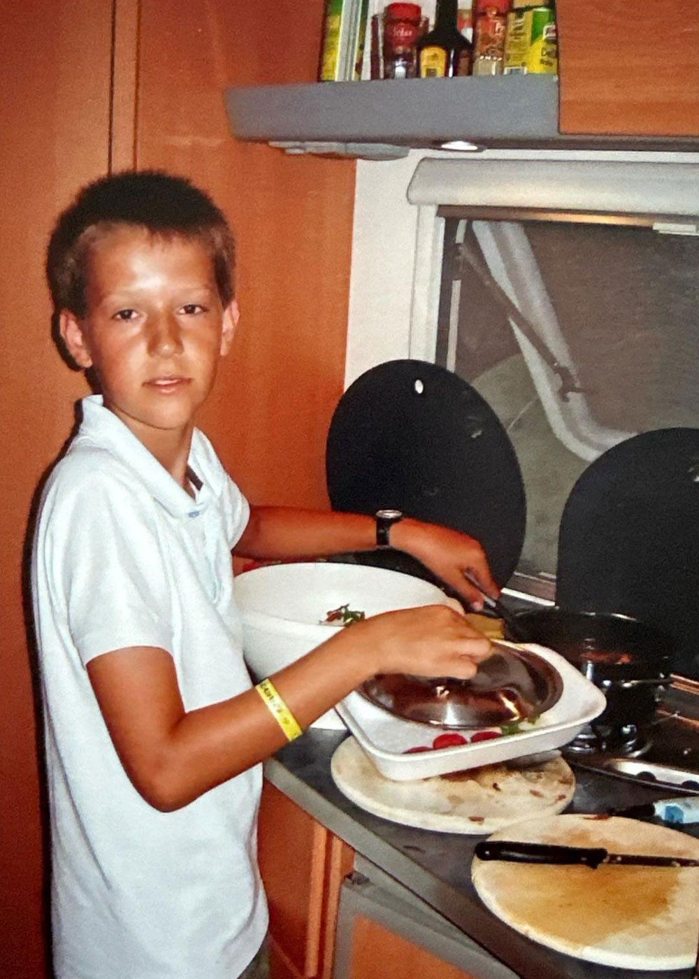 Ein altes Foto von einem kleinen Jungen. Er steht an einem Herd und kocht. Er schaut zur Kamera, hat kurze Haare und trägt ein helles T-Shirt. Im Hintergrund sind Küchenregale mit Vorräten zu sehen.