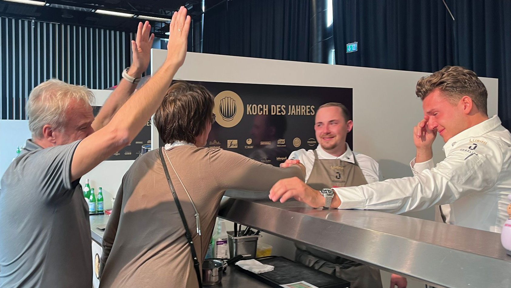 Emotionaler Moment bei der Preisverleihung «Koch des Jahres»: Vier Personen stehen um ein Metallgestell herum, eine davon jubelt mit erhobenen Armen, während ein junger Koch in weissem Kochkittel gerührt wirkt. Die Szene strahlt Freude und Erleichterung über den Erhalt einer bedeutenden Auszeichnung aus.