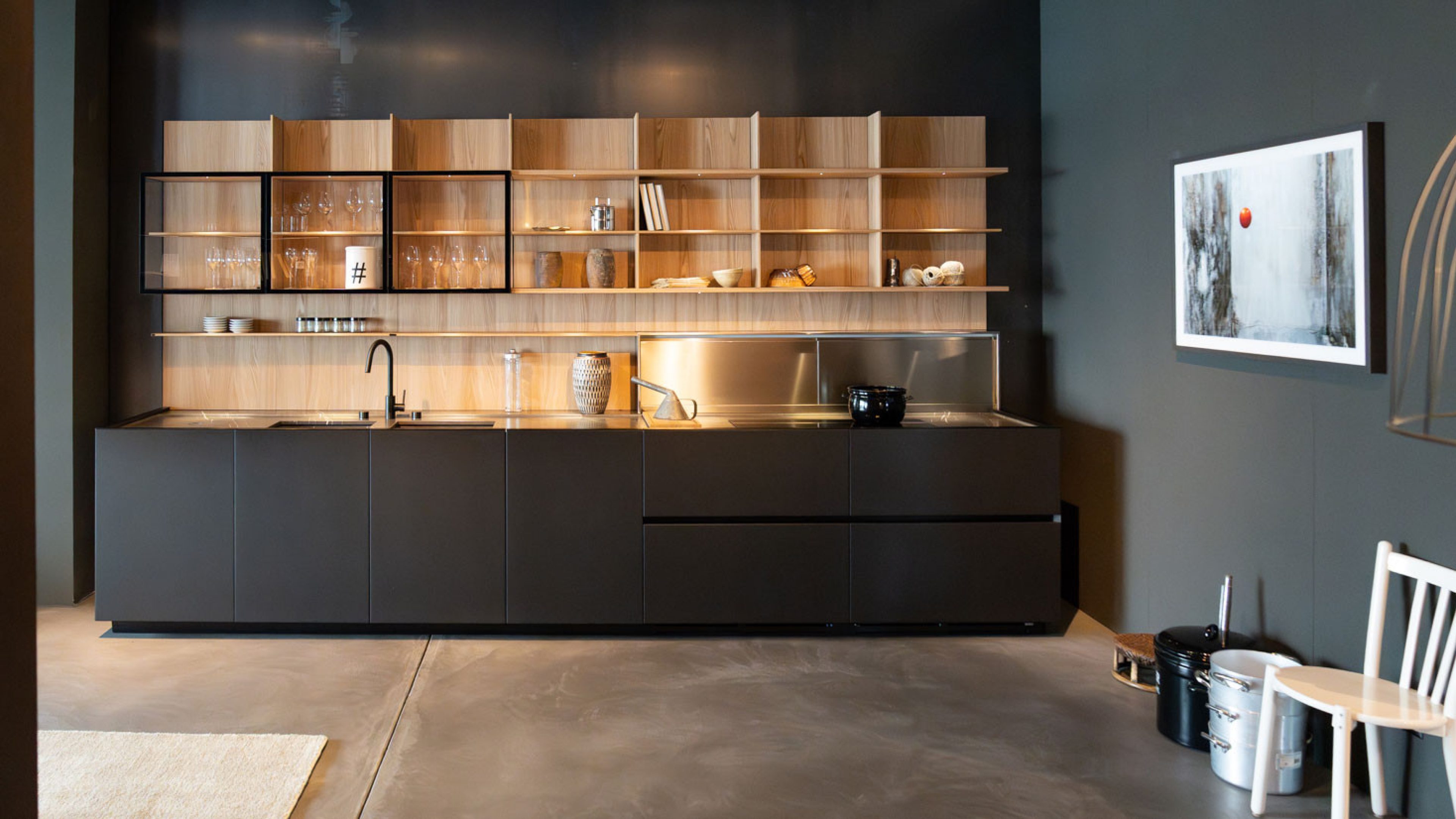 Eine moderne italienische Designerküche mit mattschwarzem Unterschrankblock, eleganter Edelstahlarbeitsfläche und offenen Holzregalen steht in einem Showroom. Die Rückwand dunkel-grau und es hängt ein eingerahmtes Bild rechts von der Küche. Gläser, Tassen und Dekoration sind stilvoll arrangiert.
