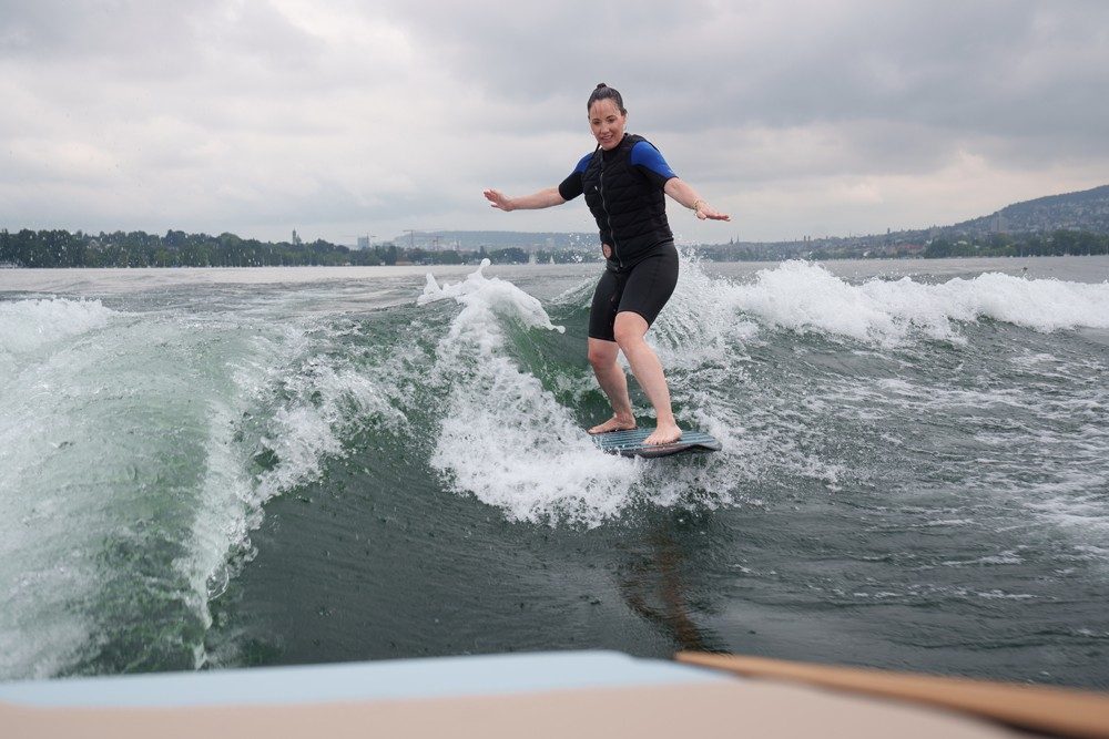 Eine Frau in einem schwarzen Neoprenanzug reitet auf einem schwarzen Wakesurf-Brett eine vom Schiff ausgehende Wasserwelle. Sie balanciert mit ihren Armen und sieht glücklich aus.