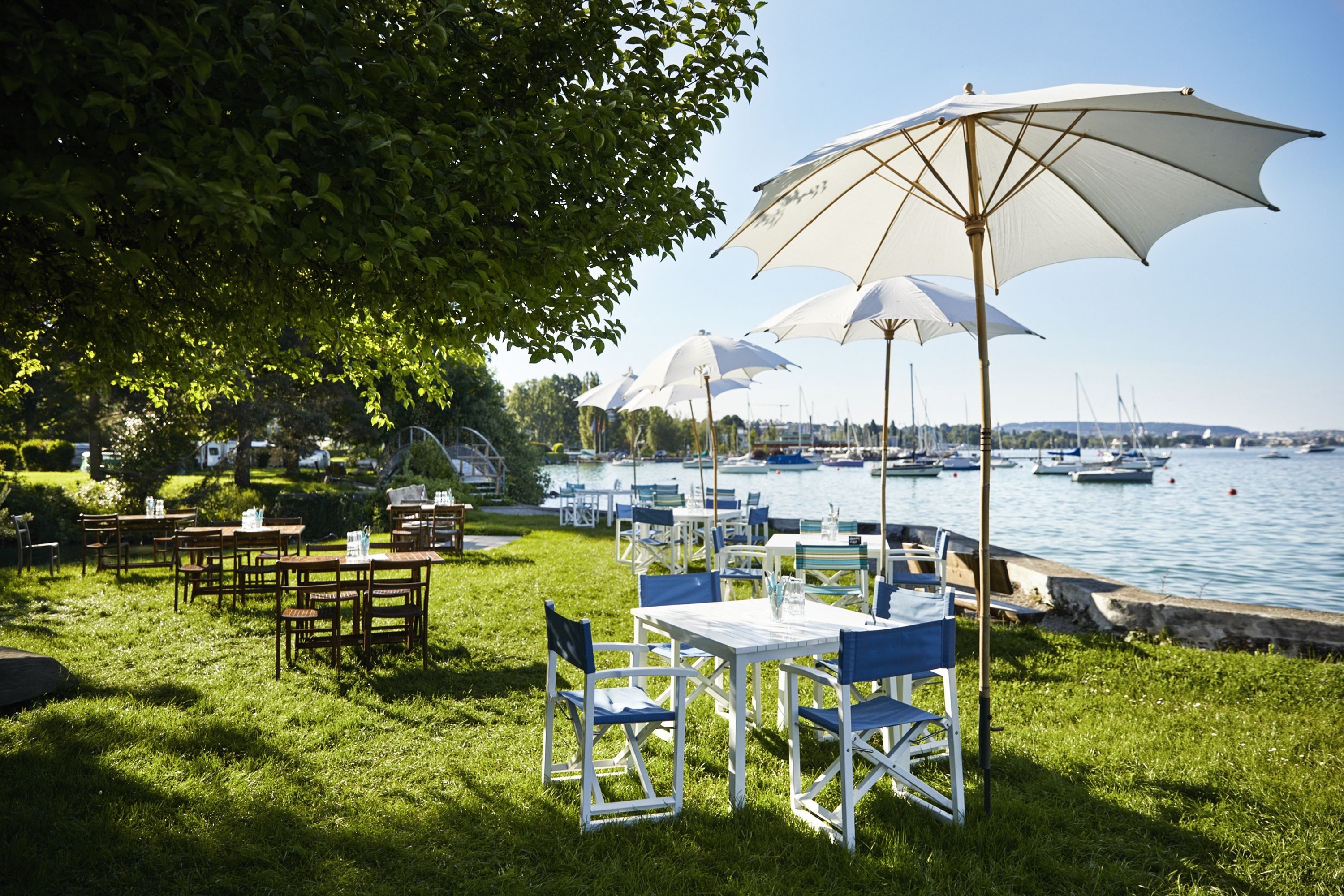 Auf einer Wiese neben einem Seeufer stehen Restauranttische mit weissen Sonnenschirmen. 