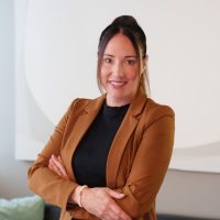 Eine Frau blickt freundlich lachend mit verschränkten Armen in die Kamera. Sie trägt einen braunen Blazer und einen schwarzen Pullover. Hinter ihr sind eine Couch und eine Leinwand.