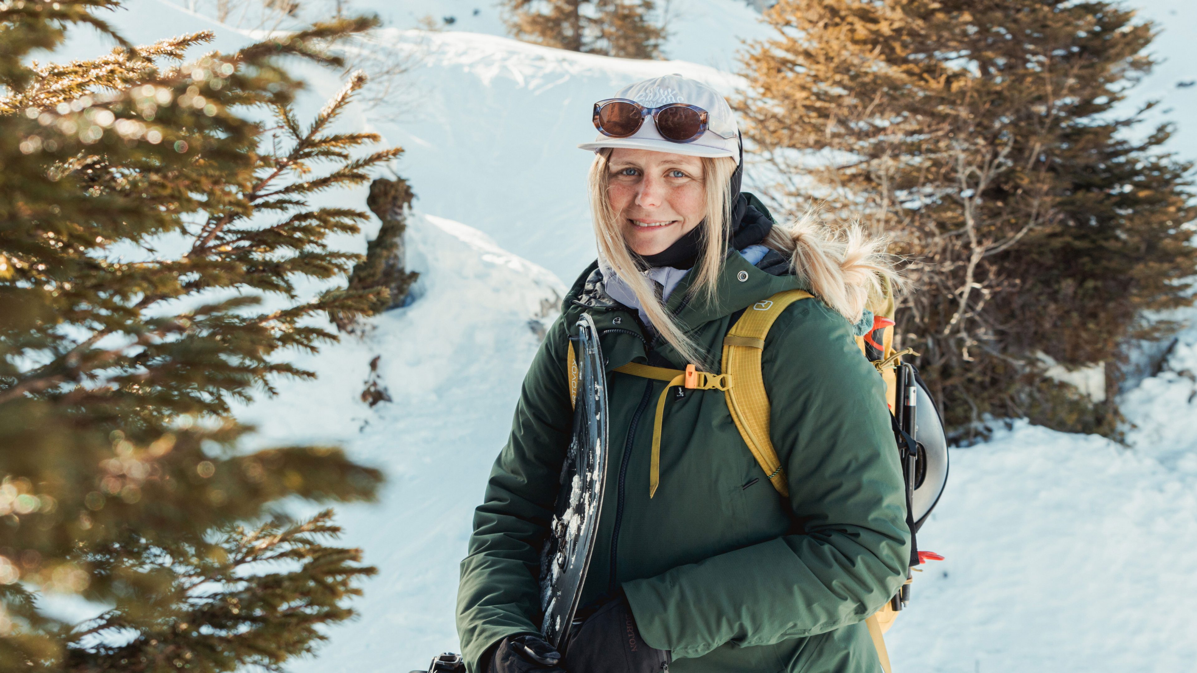 Person trägt ein Snowboard und wandert durch eine verschneite Berglandschaft zwischen winterlichen Nadelbäumen. 