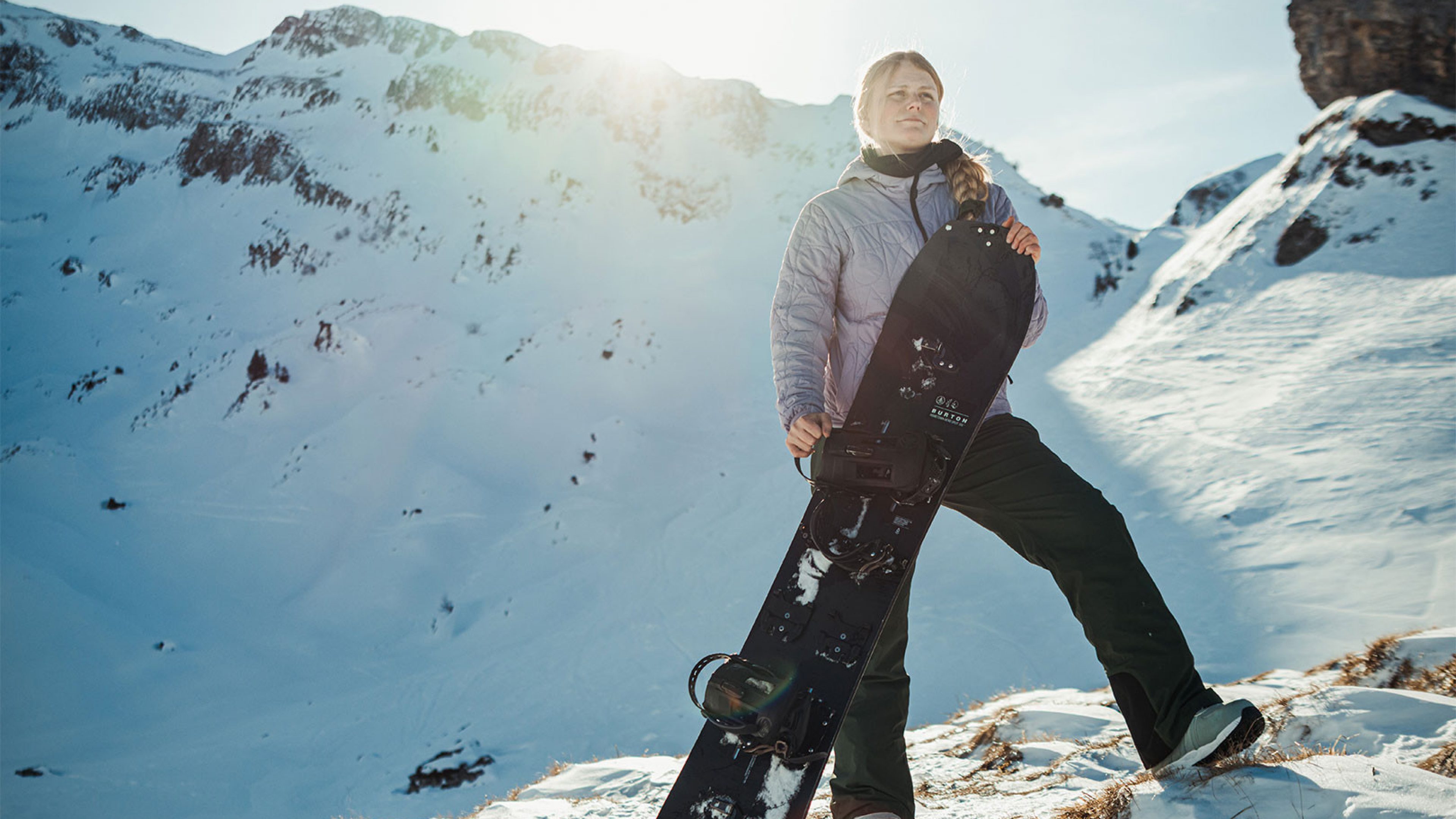Person mit Snowboard steht auf einem verschneiten Berghang in der Sonne, umgeben von alpiner Winterlandschaft. 
