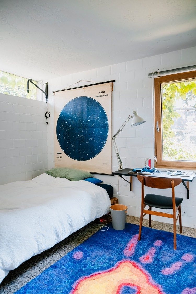 In der Ecke eines weissen, lichtdurchfluteten Zimmers steht ein kleines Bett, über dem eine Sternenkarte hängt. Neben dem Bett steht ein Arbeitstisch mit einem Holzstuhl auf einem Teppich mit einem Thermo-Handabdruck-Motiv.