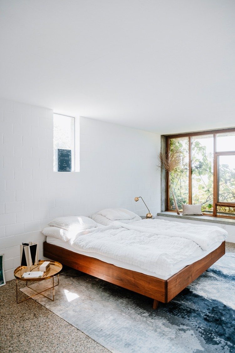 Ein Bett mit einem Holzrahmen steht auf einem blau-weissen Teppich, der an einen eisigen Fluss erinnert. Neben dem Bett ist auf einer Seite ein kleiner bronzefarbener Abstelltisch und auf der anderen ein grosses Fenster, das Sicht auf Bäume gewährt.