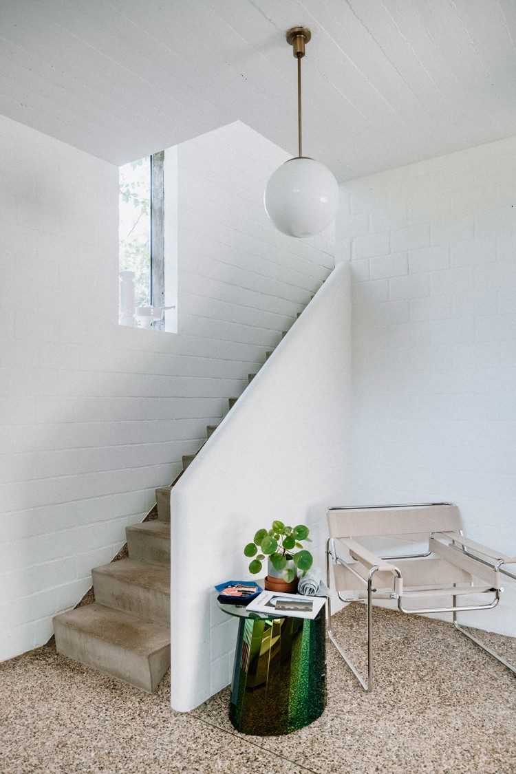 Auf einem Waschbetonboden stehen ein Stuhl und ein grüner Beistelltisch mit einer Pflanze. Hinter dem Sitzplatz ist eine Treppe. Die Wände und die runde Lampe sind weiss.