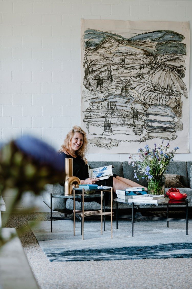 Eine blonde Frau in einem dunkelblauen Pullover sitzt mit einem Buch in der Hand auf einem grauen Sofa und lächelt in die Kamera. Hinter ihr hängt eine grosse Kohlezeichnung und um sie herum stehen Vasen mit blauen Blumen und Bücherstapeln.