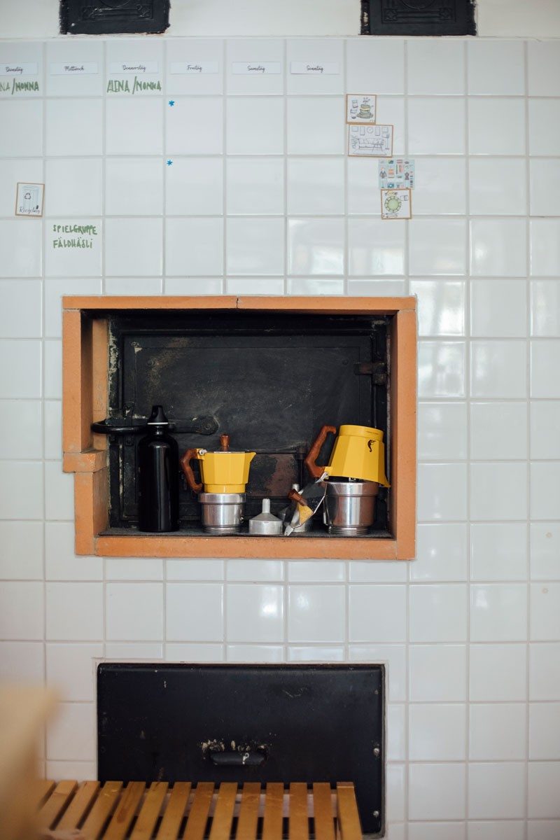 Eine geflieste Wand mit einem eingebauten Ofen, davor stehen Utensilien zum Kaffeekochen.