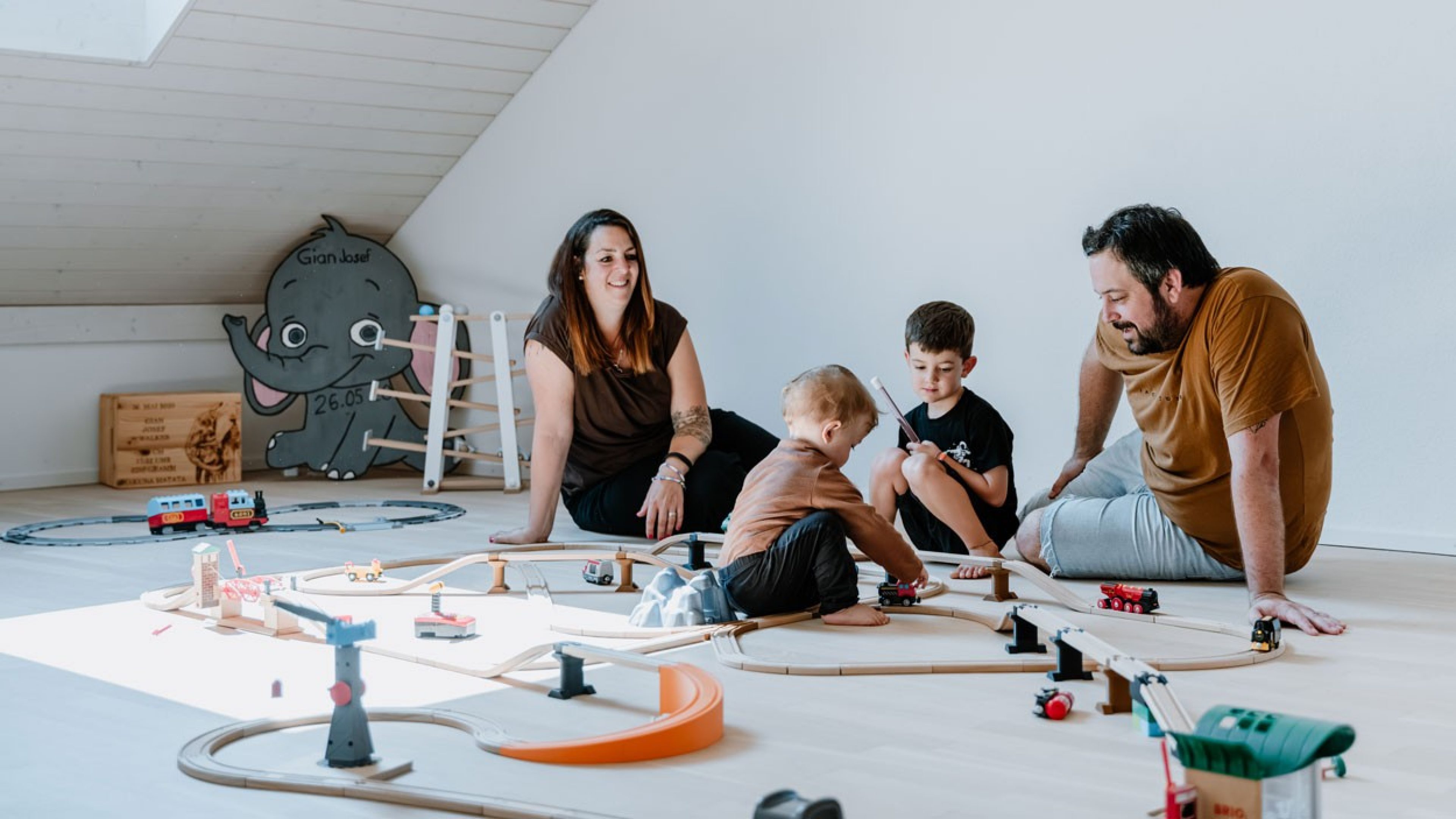 Eine Frau mit braunen Haaren und einem Tattoo am Ellbogen und ein Mann mit braunen Haaren und einem braunen T-Shirt spielen mit zwei Kindern am Boden eines Dachzimmers. Auf dem Boden liegt verschiedenes Spielzeug. Die Szene zeigt eine warme, familiäre Atmosphäre.